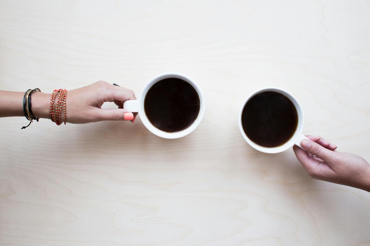 Two female hands holding coffee mugs on a light wooden table, symbolizing friendship and relaxation.