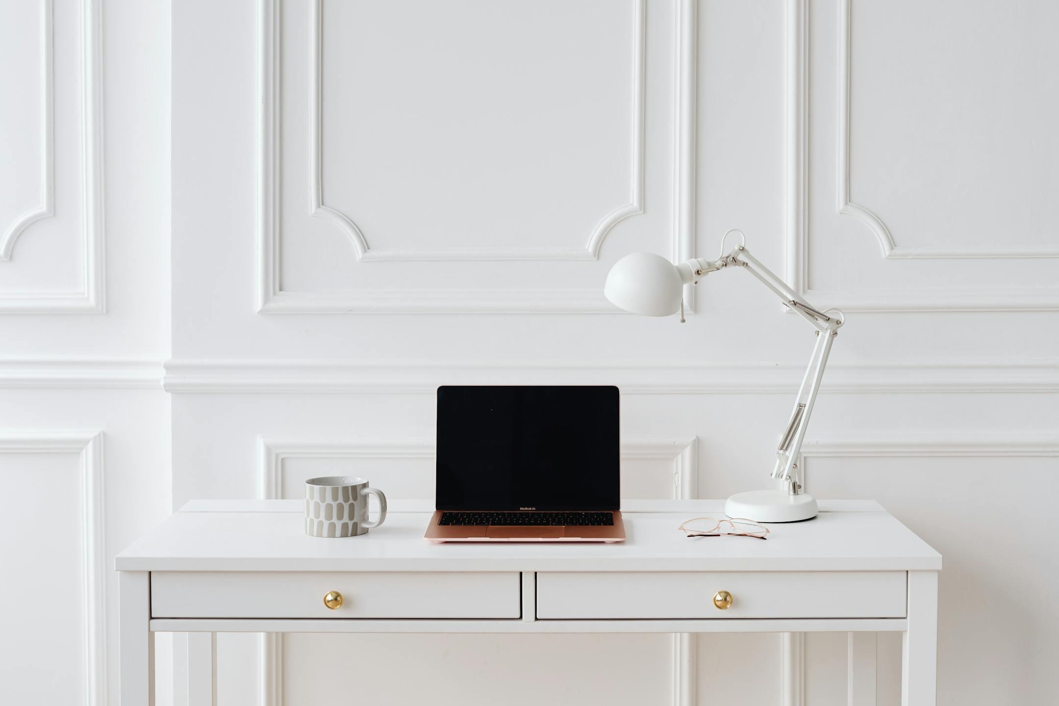 Elegant minimalist workspace with laptop and desk lamp on a white desk in a bright room.