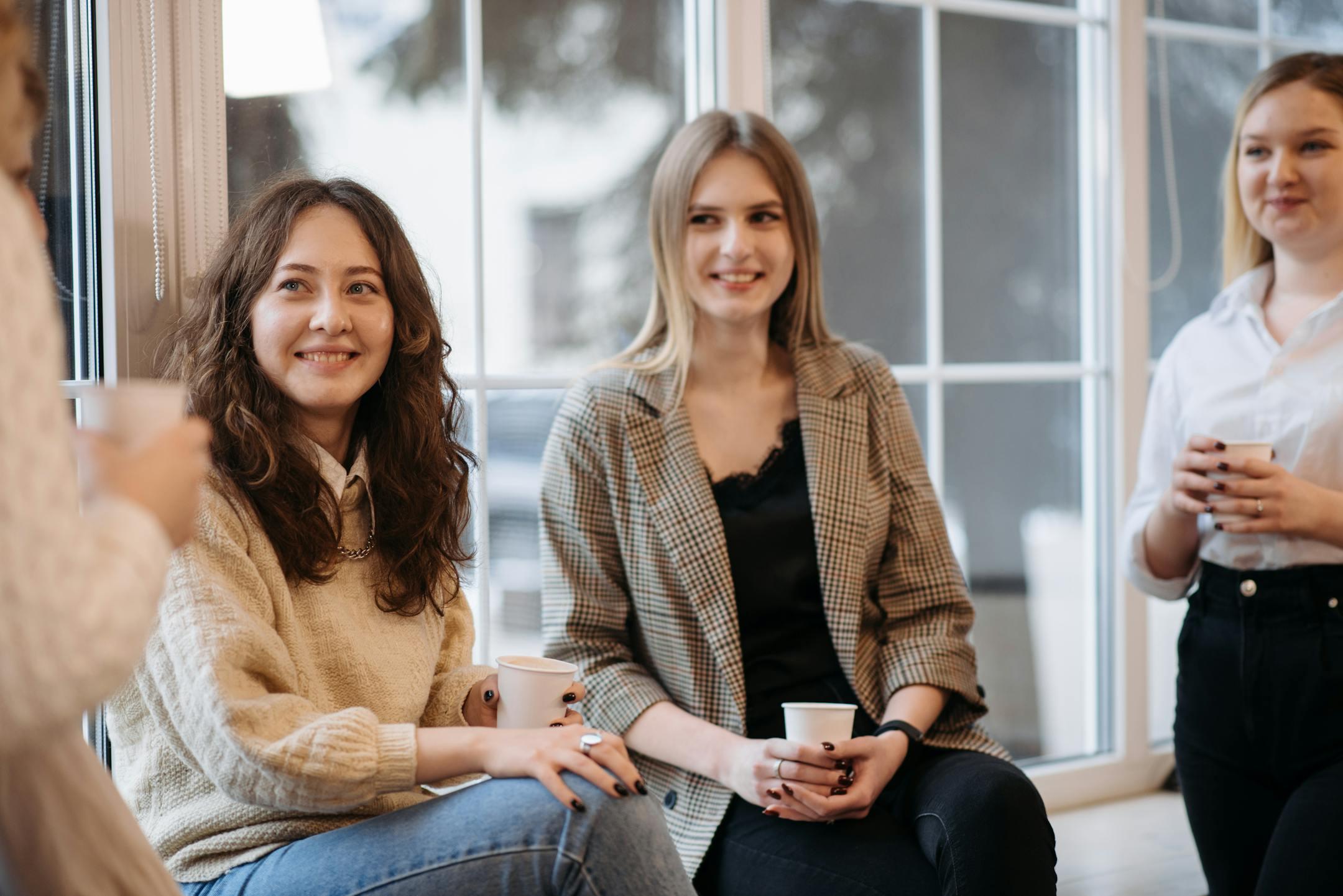 Three women in casual attire enjoy a friendly coffee break indoors by the window.