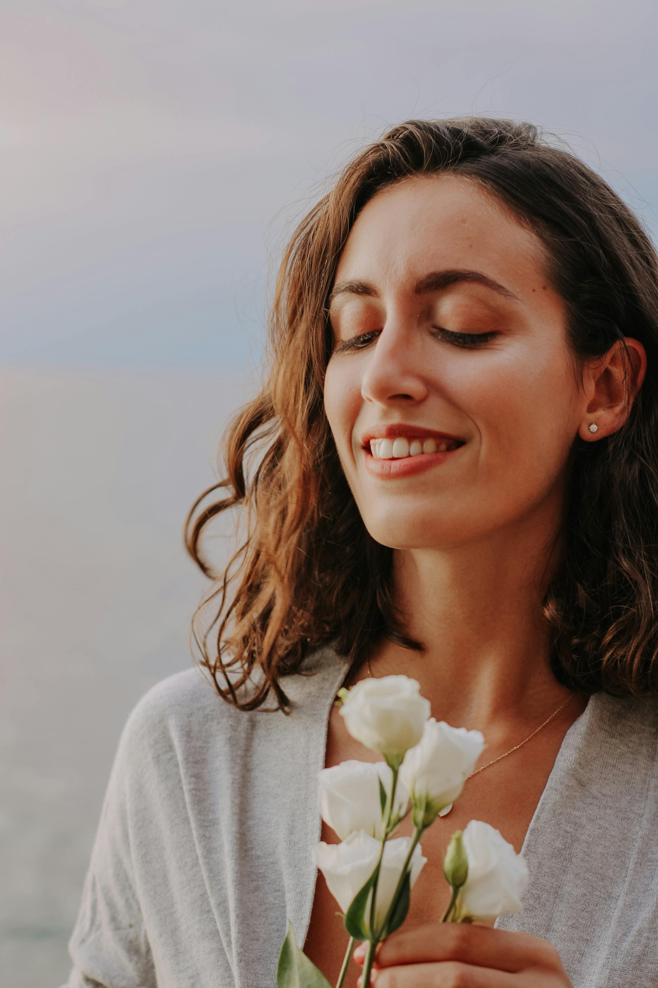 A serene woman enjoying a moment by the sea, holding white roses with eyes closed and smiling.