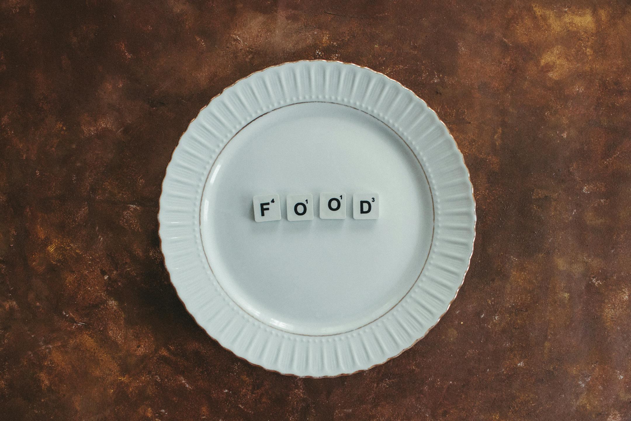 A minimalist composition featuring a white plate with the word 'food' in scrabble tiles on a textured background.