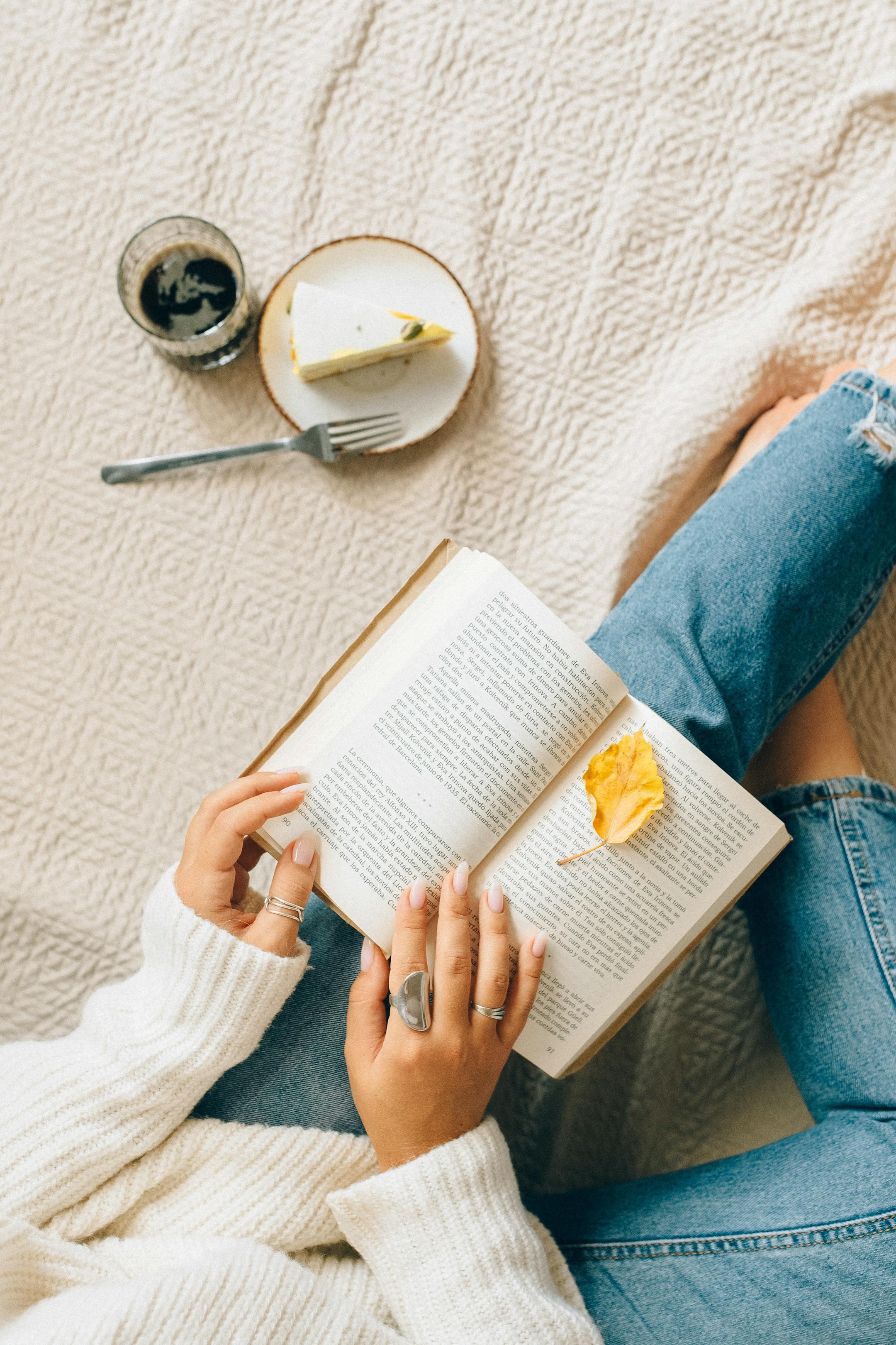 Woman enjoying leisure time at home with a book, coffee, and cheesecake on a cozy blanket.