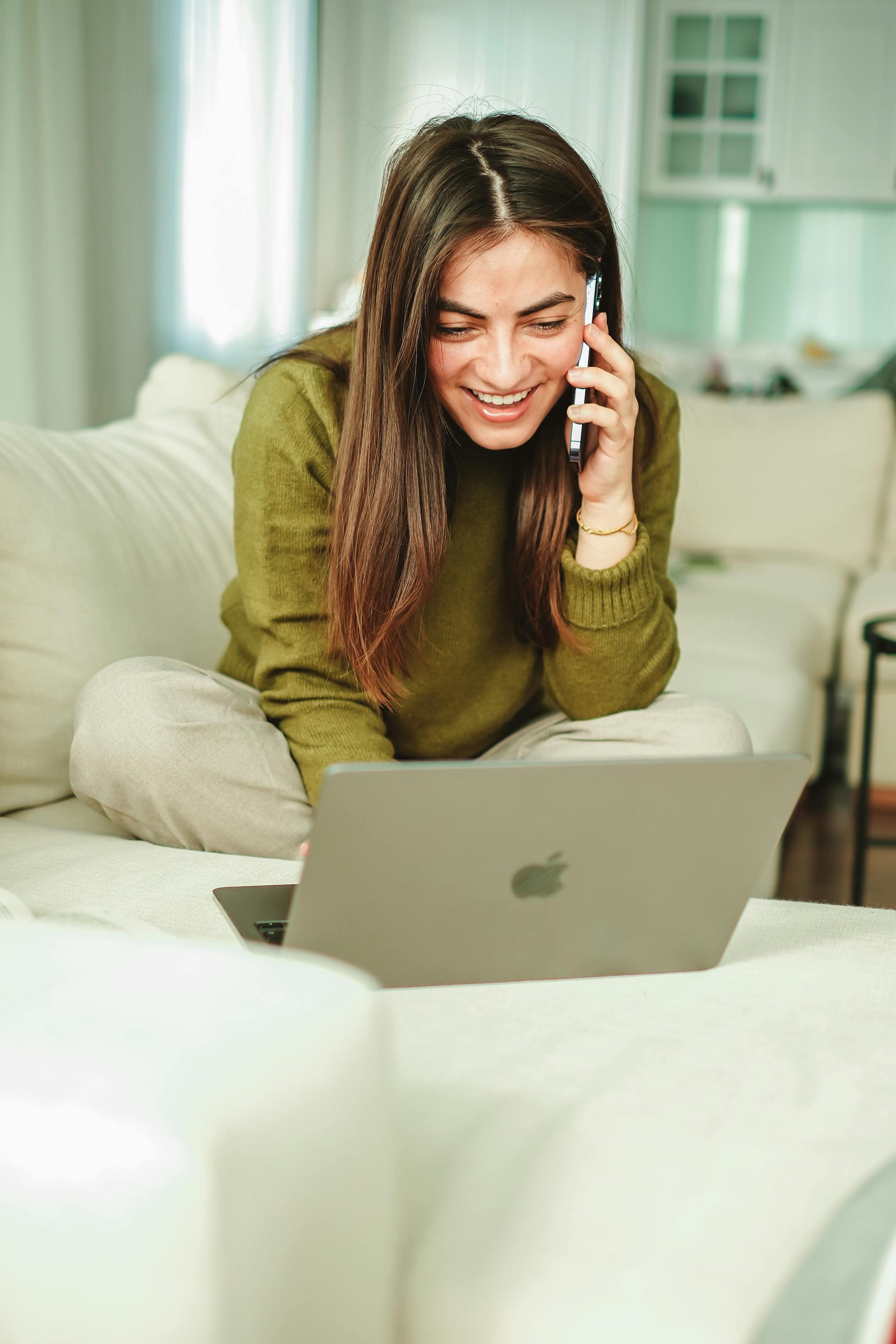 A woman sits on a sofa, smiling while talking on a smartphone and using a laptop, embodying remote work lifestyle.