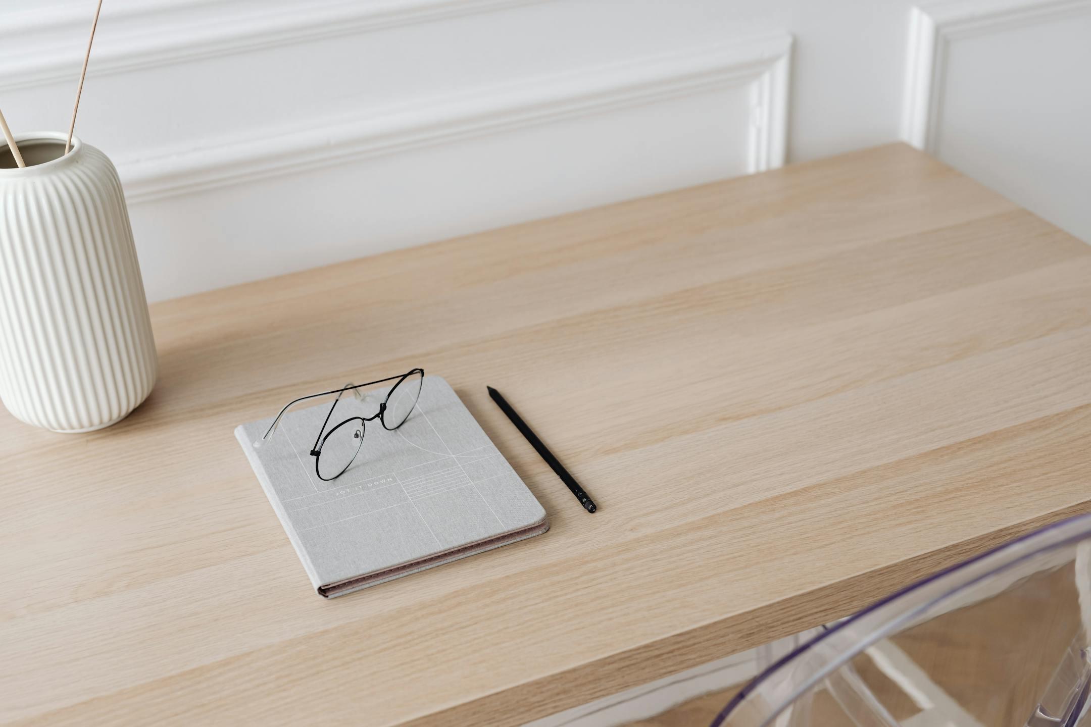 A minimalist workspace featuring a notebook, eyeglasses, pen, and a decorative vase on a wooden desk.