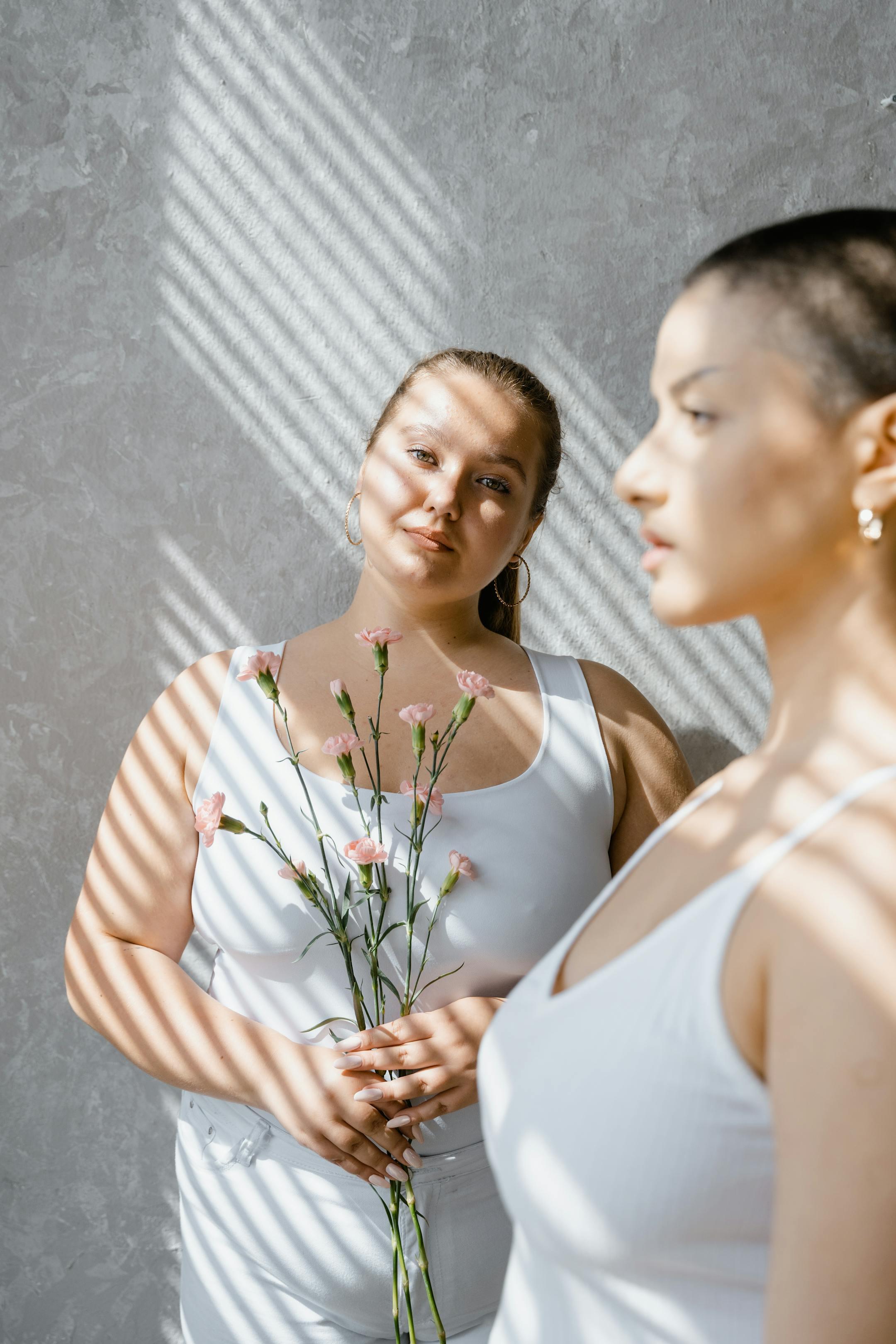 Two women in white attire standing against a textured wall with floral accents.