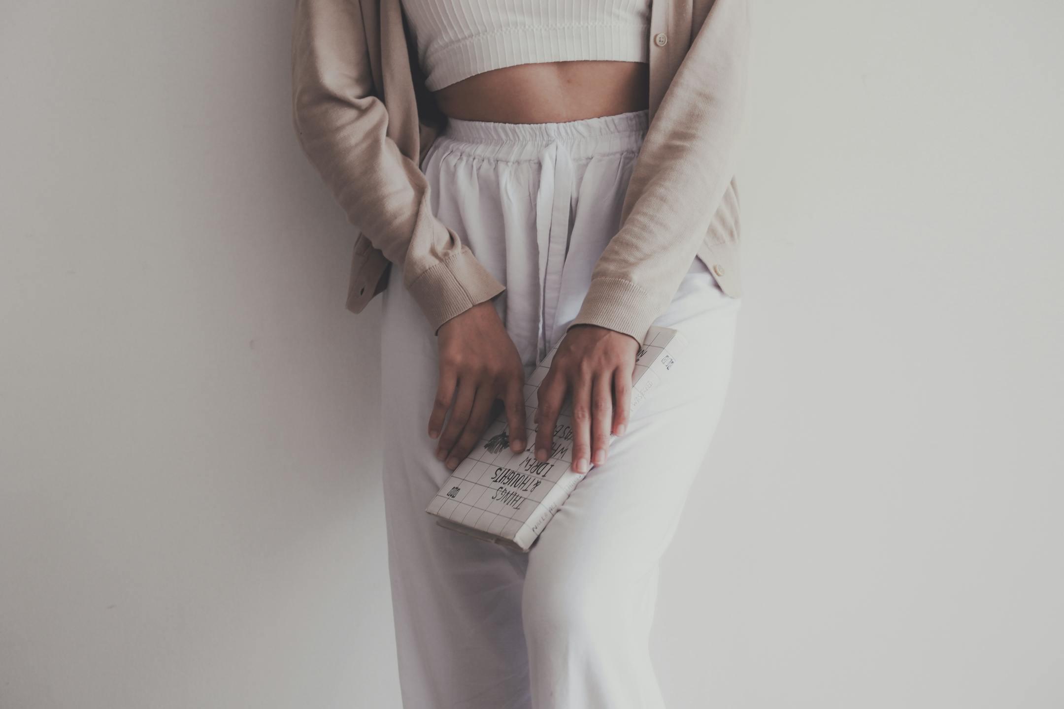 Stylish woman in beige cardigan and white crop top holding a book against a white background.