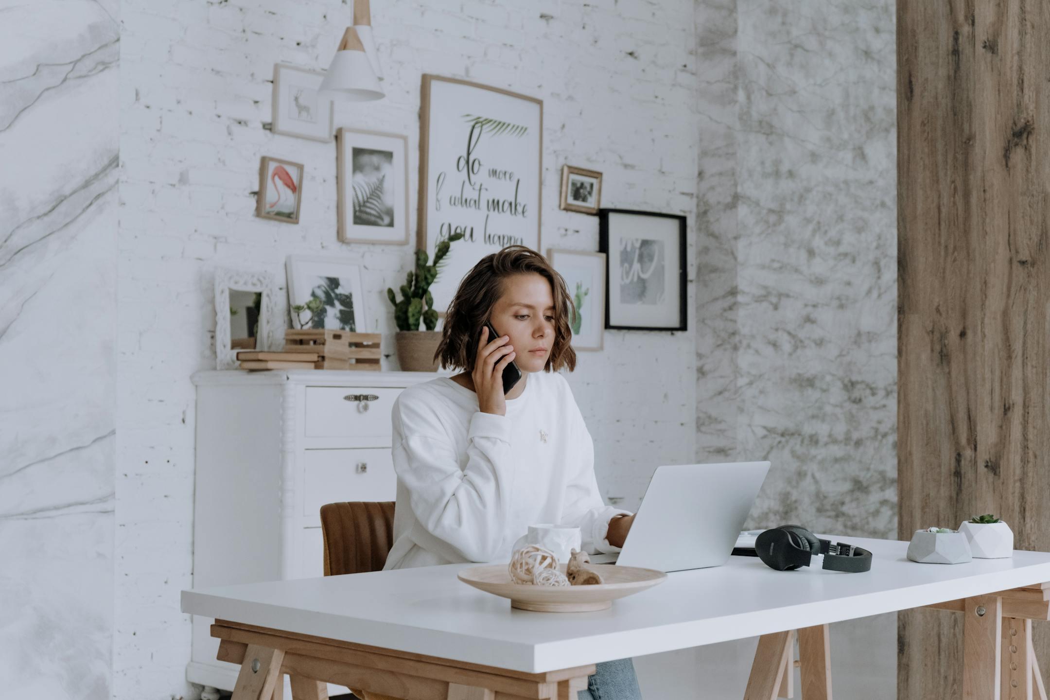 Woman talking on phone while using laptop in bright, modern home office with decor.