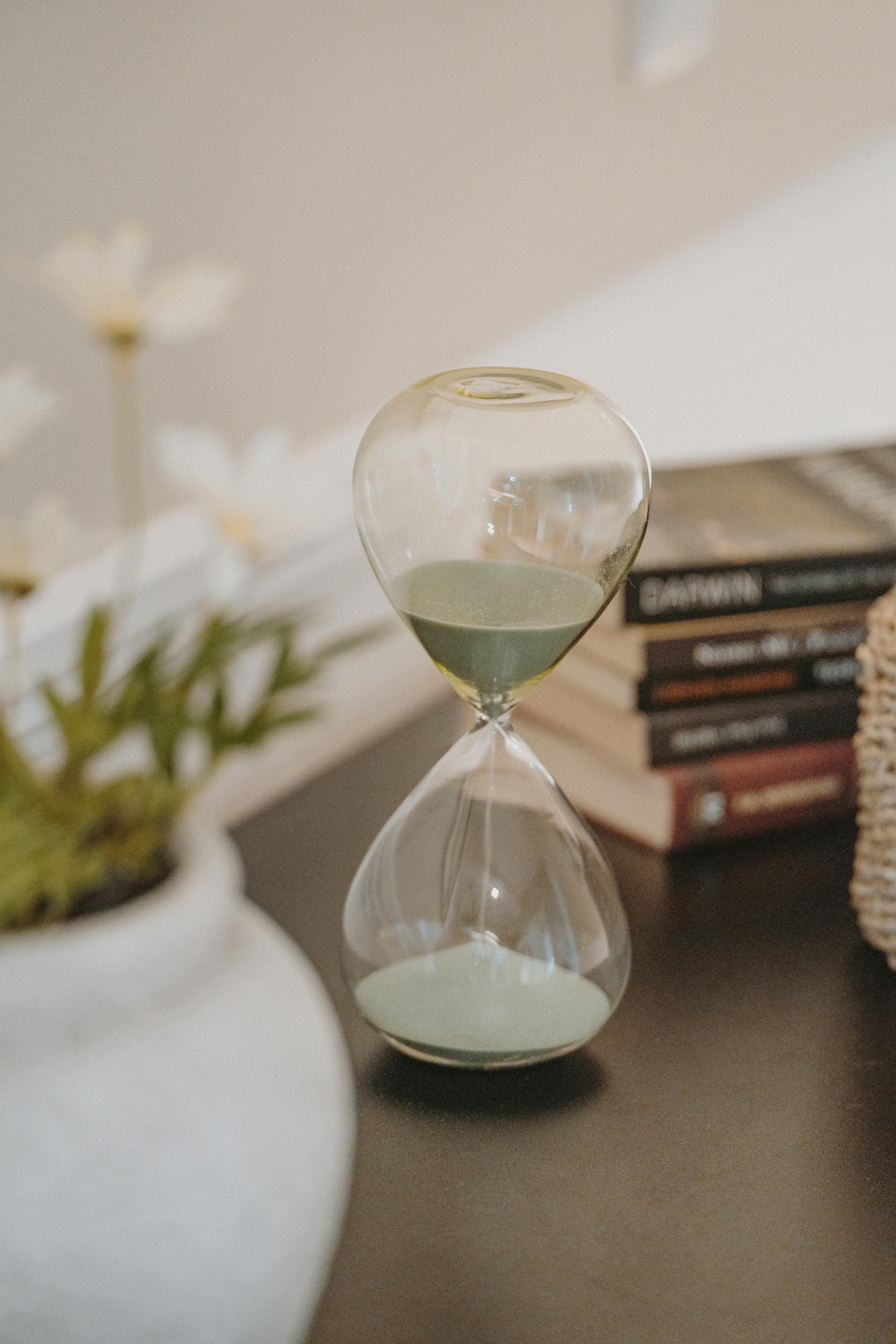 Stylish hourglass with green sand beside books and decor on table in elegant setting.