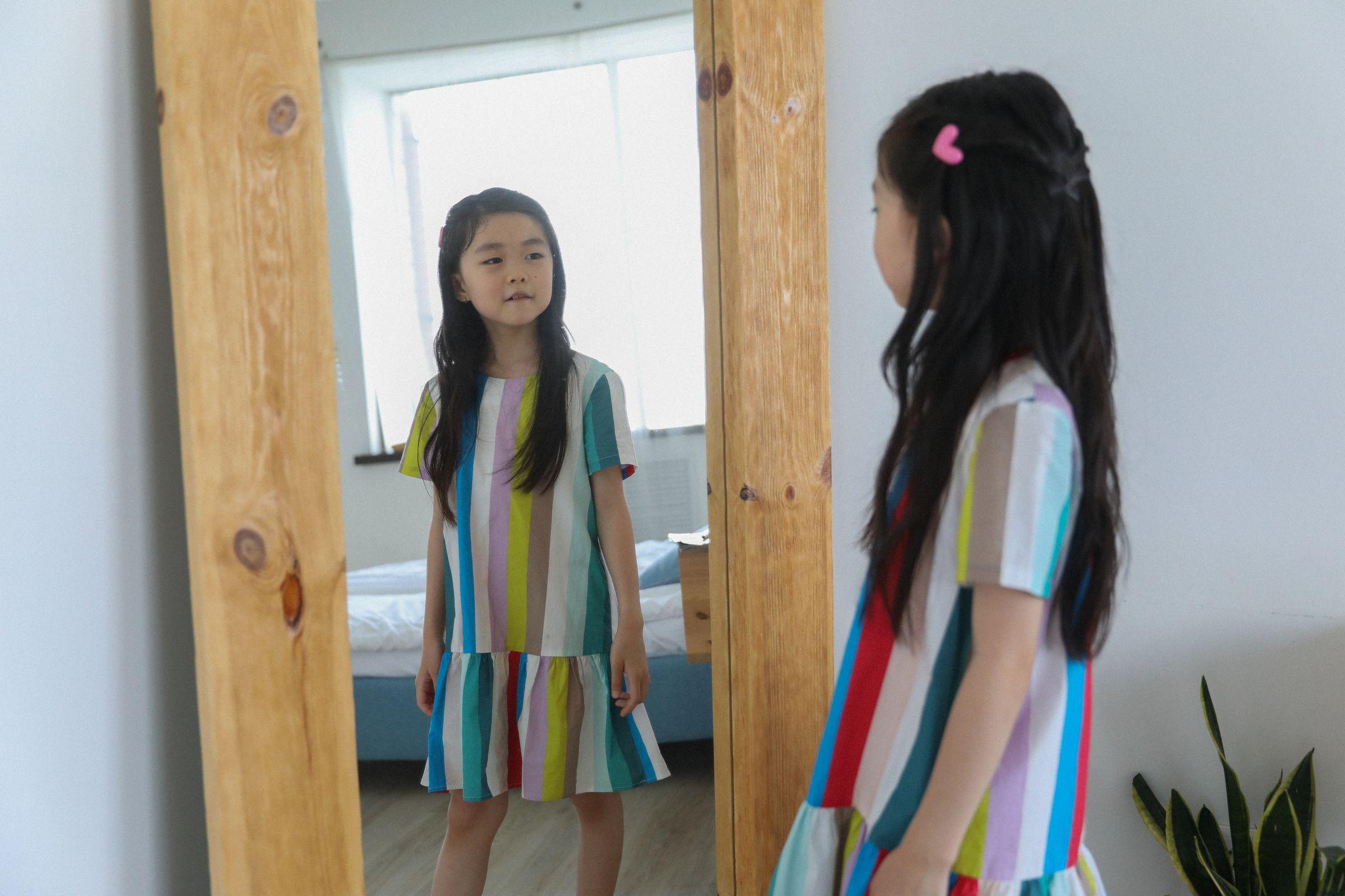 Cute Asian girl in bright dress with colorful stripes reflecting in mirror with wooden frame