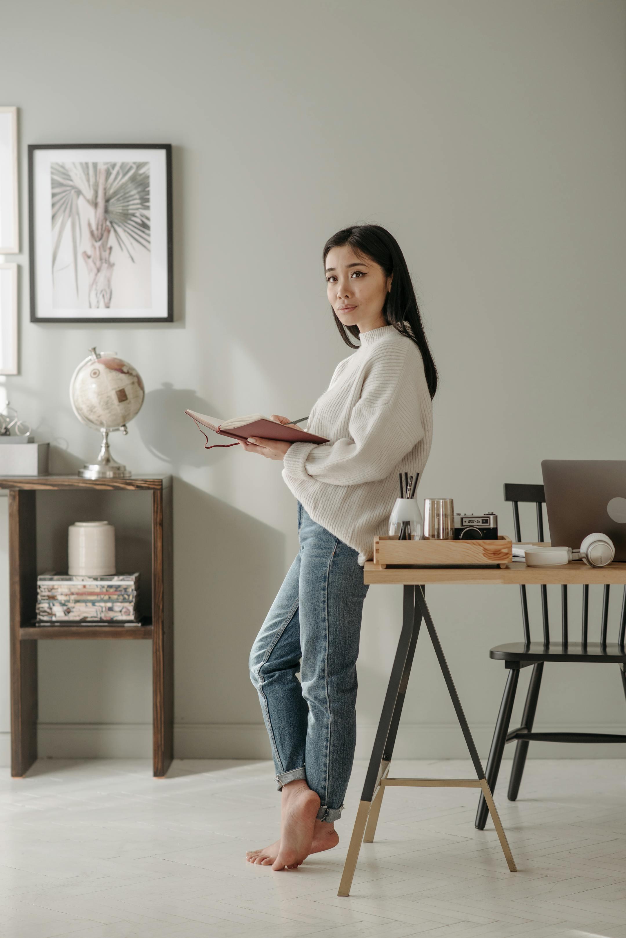Asian woman in a cozy setting holding a book, contemplating by the table.