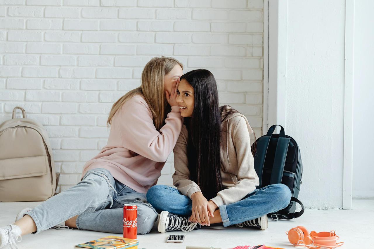 Two teenage girls whispering and sharing secrets while sitting indoors, surrounded by backpacks.
