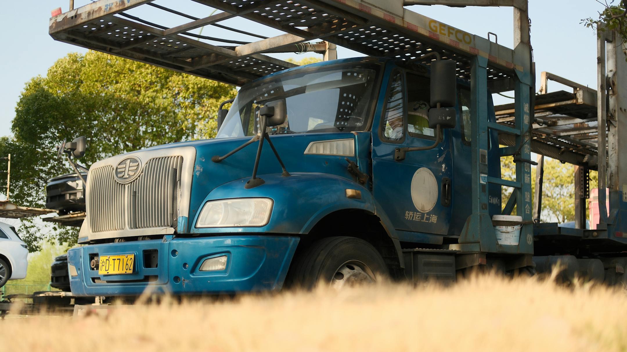 Blue car transport truck parked outdoors with tree background.