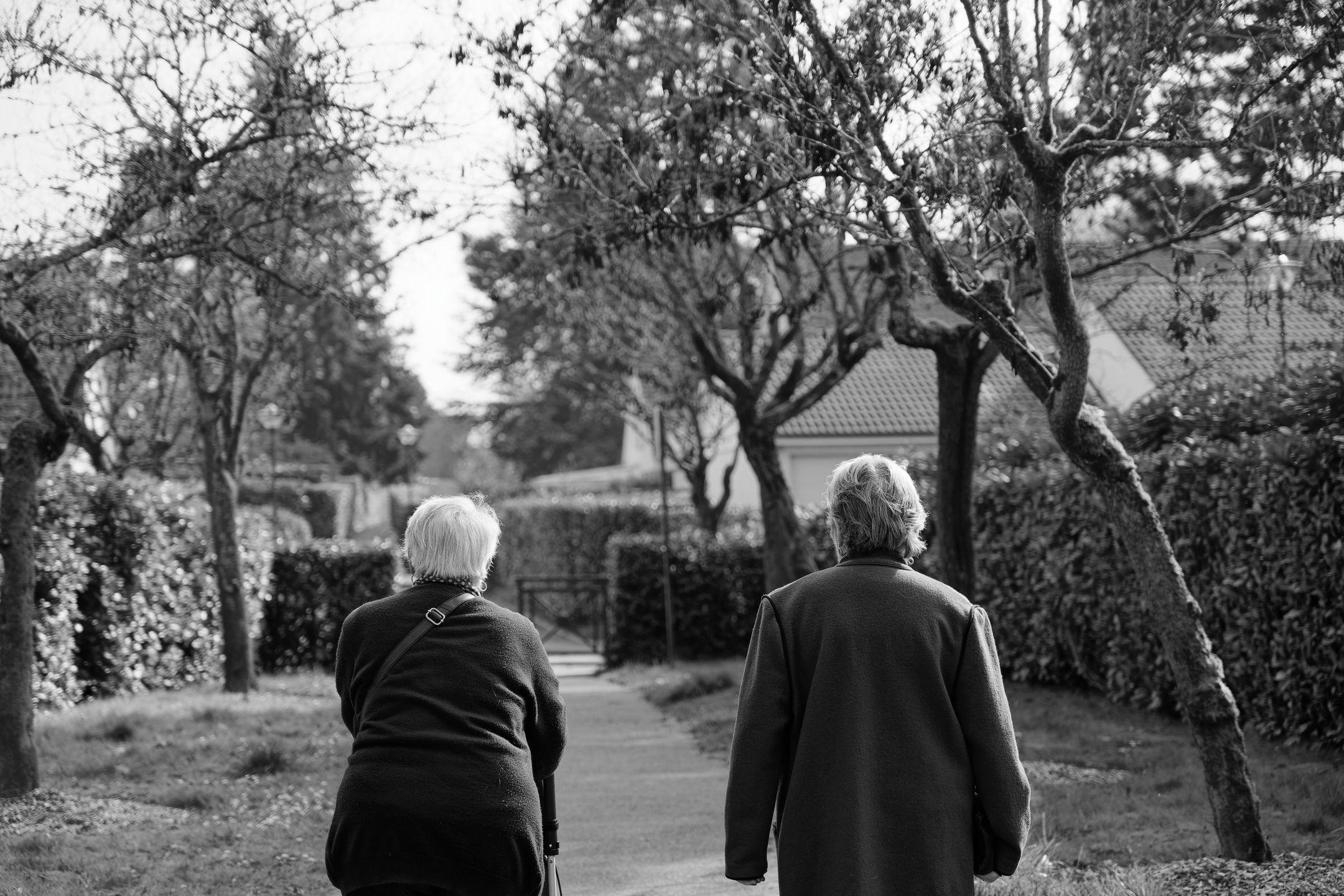 Black and white photo of two senior adults walking through a park during the day.