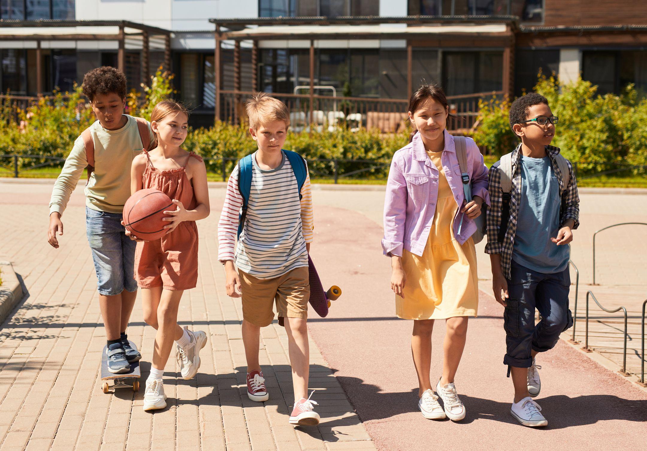 A diverse group of children walking outdoors, carrying backpacks and a basketball, heading to school on a sunny day.