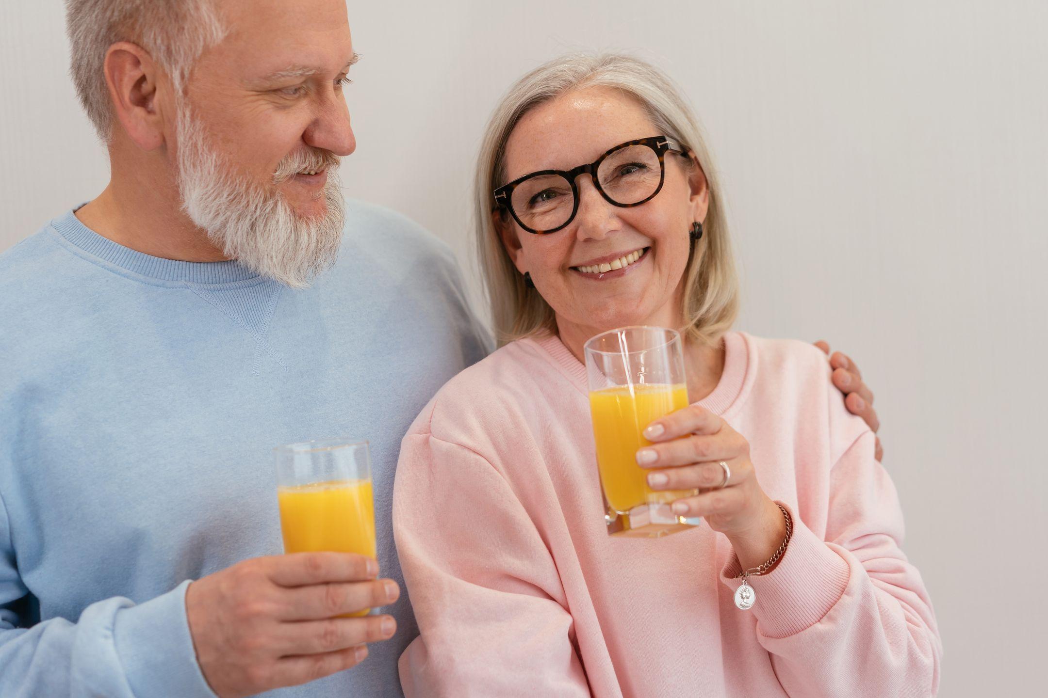 Smiling elderly couple holding glasses of fresh orange juice indoors.