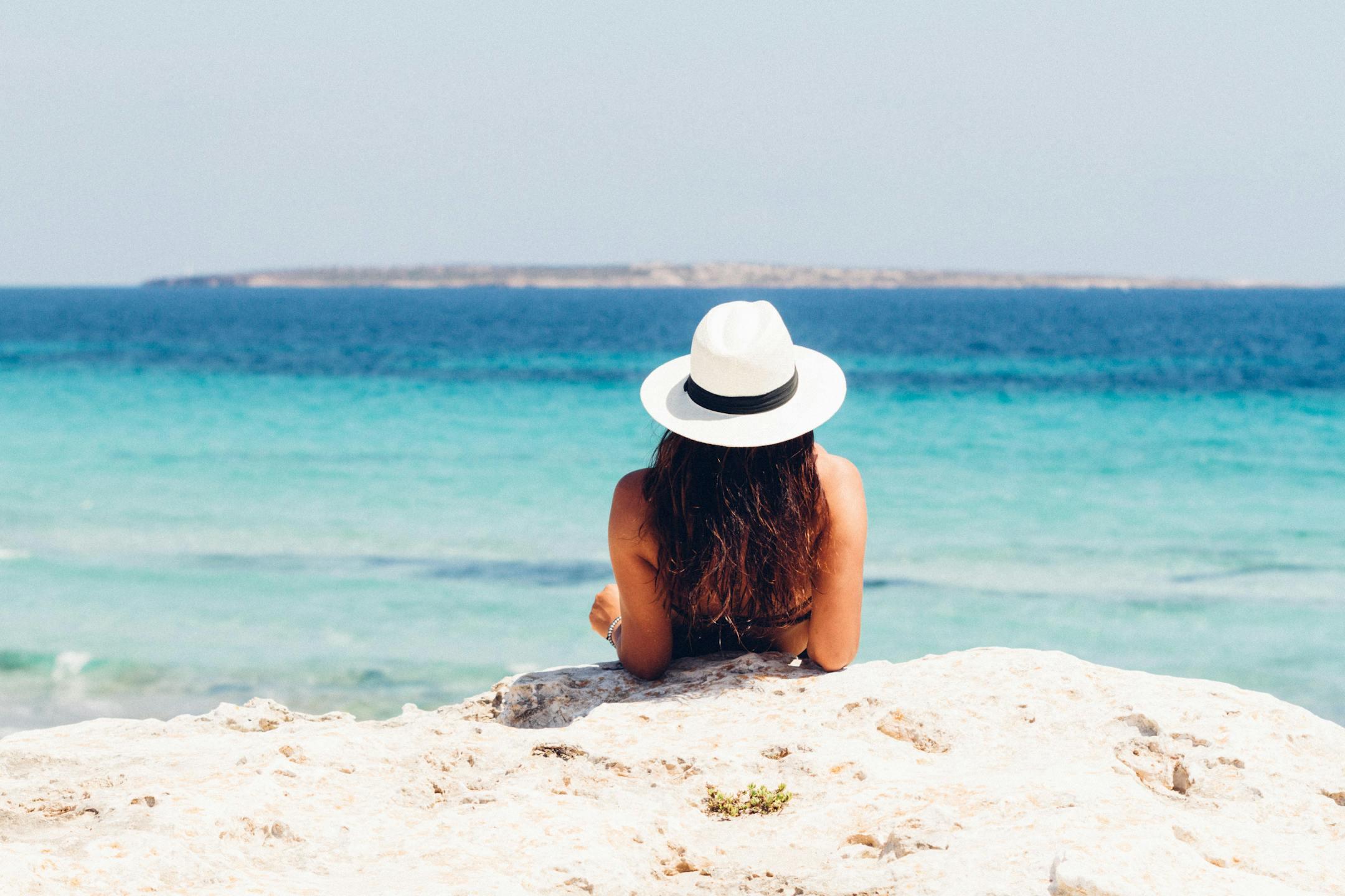 Back view of a woman in a hat relaxing on a rocky beach, facing the clear blue sea.