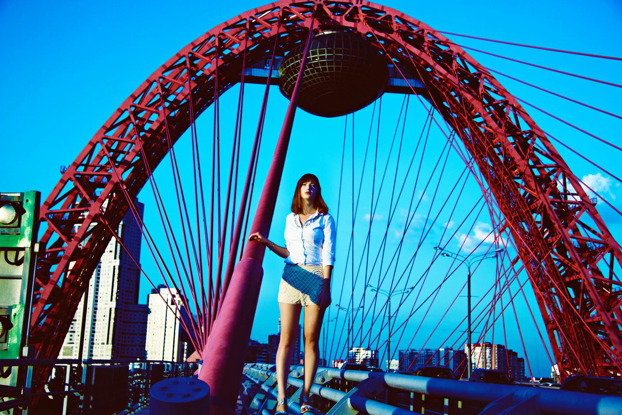 A fashionable woman poses on the Zhivopisny Bridge in Moscow, showcasing urban style against a vivid backdrop.