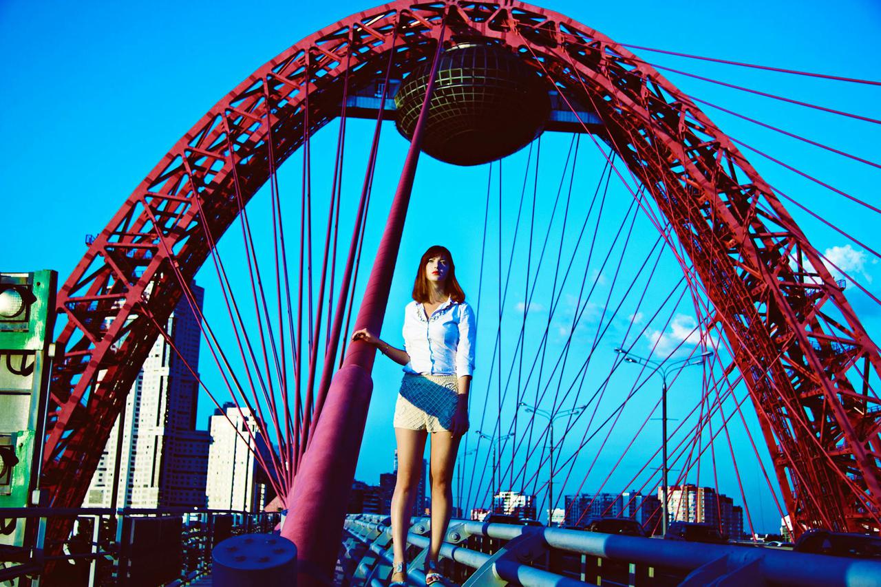 A fashionable woman poses on the Zhivopisny Bridge in Moscow, showcasing urban style against a vivid backdrop.
