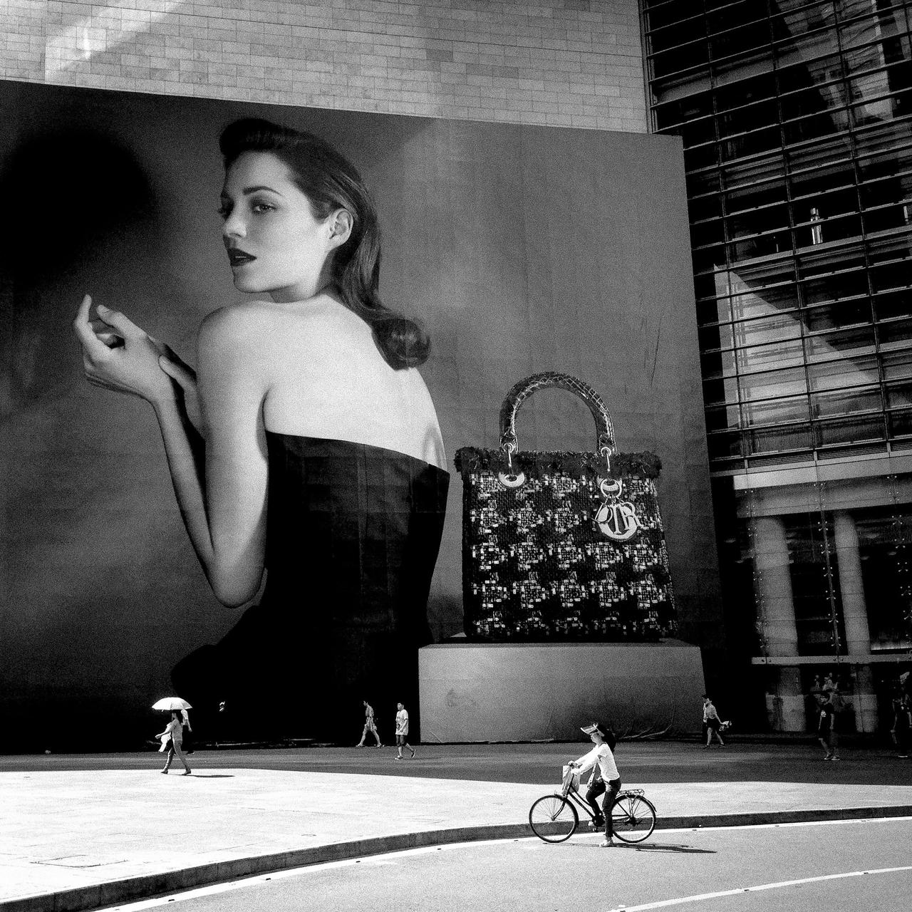 Black and white city street with a fashion ad billboard, cyclist, and pedestrians.