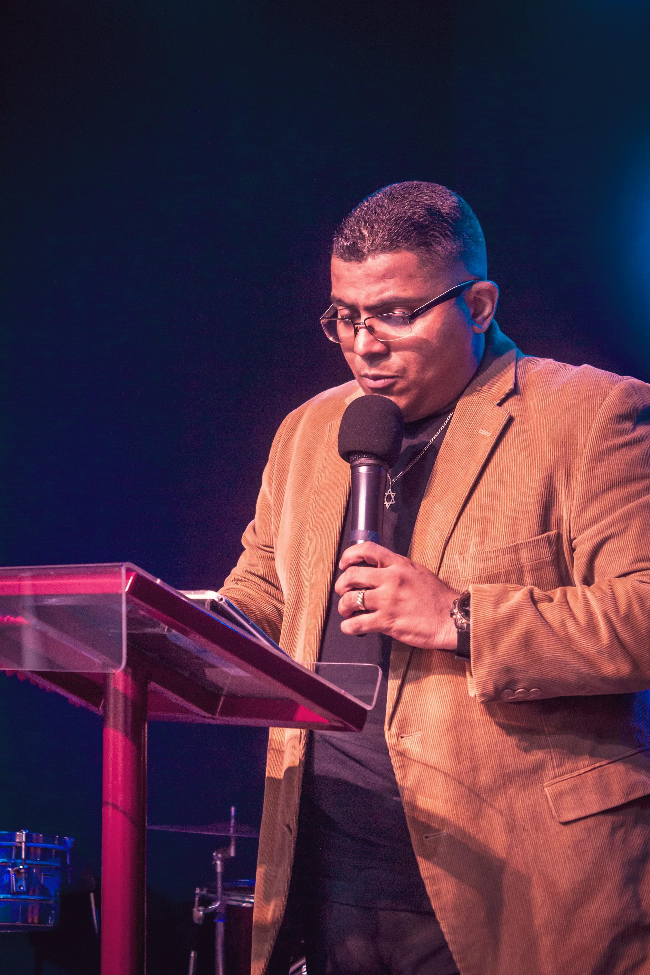 A confident man in a brown blazer delivering a speech at a podium with a microphone.