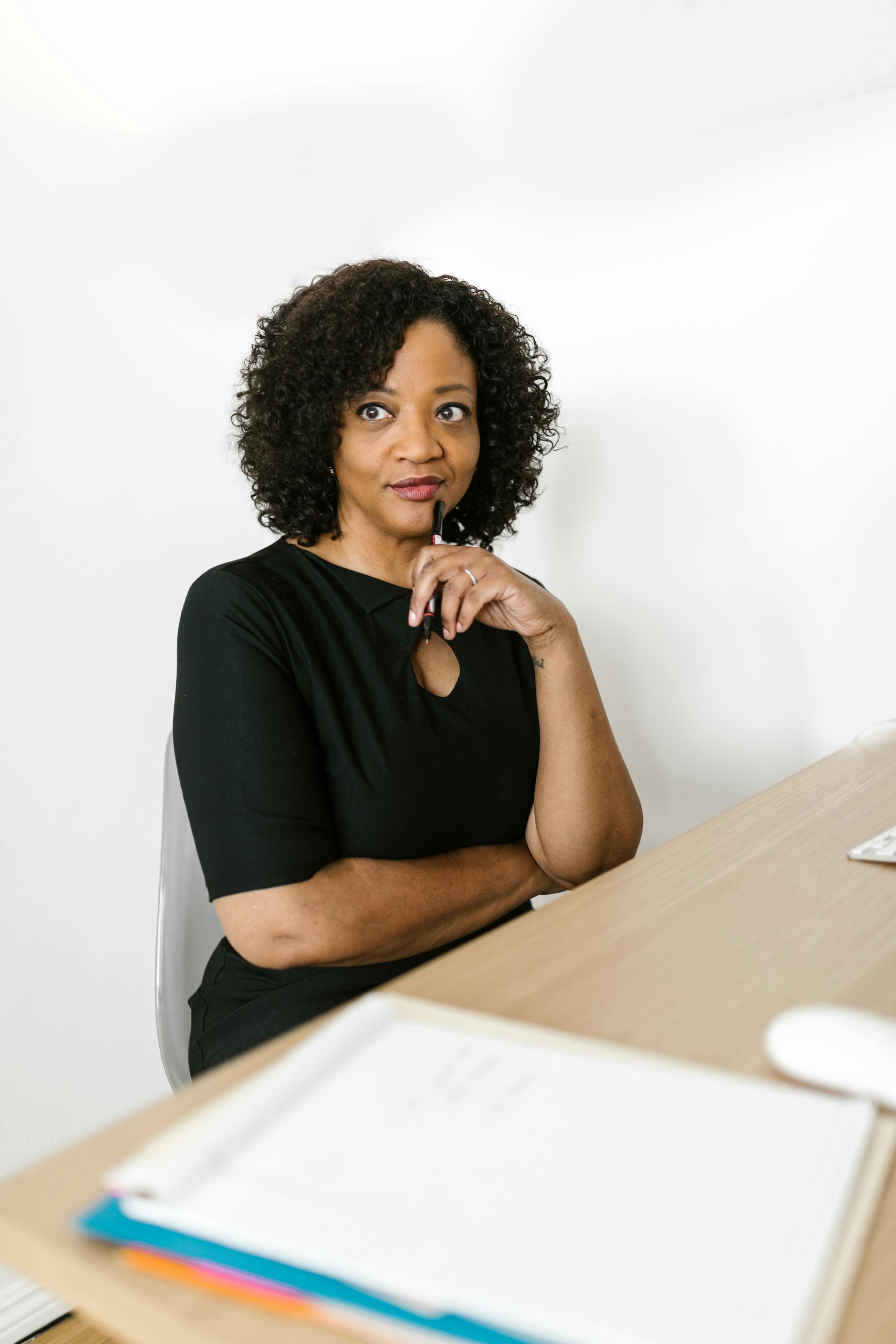 African American woman in a black dress sitting at a desk with a thoughtful expression.