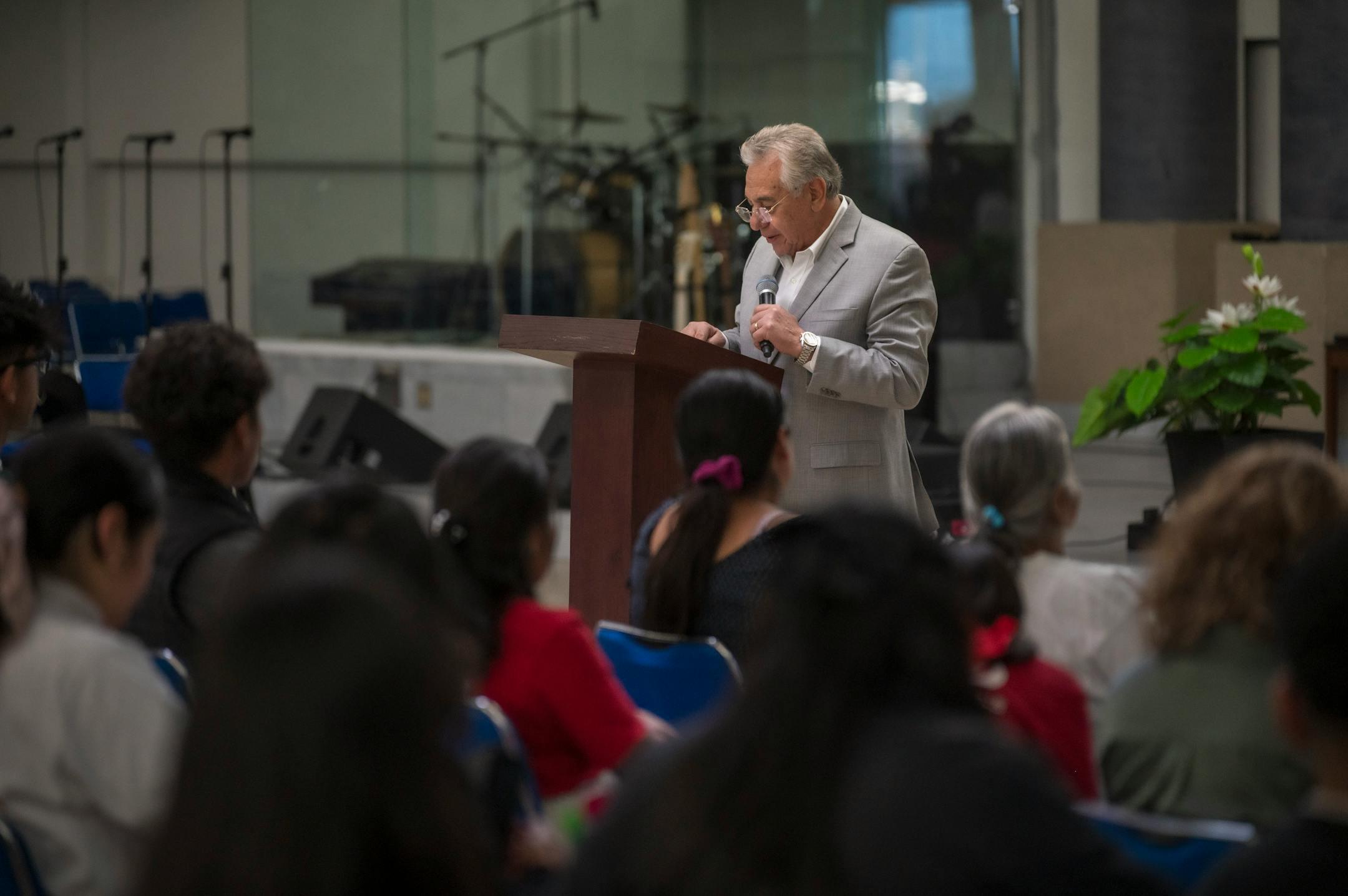 An elderly man in a gray suit delivers a speech to a seated audience indoors.