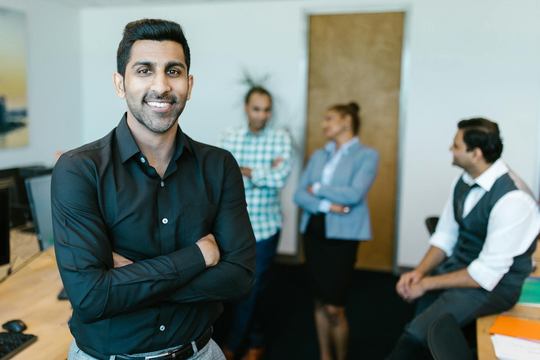 A confident young professional standing in a modern office with colleagues interacting in the background.