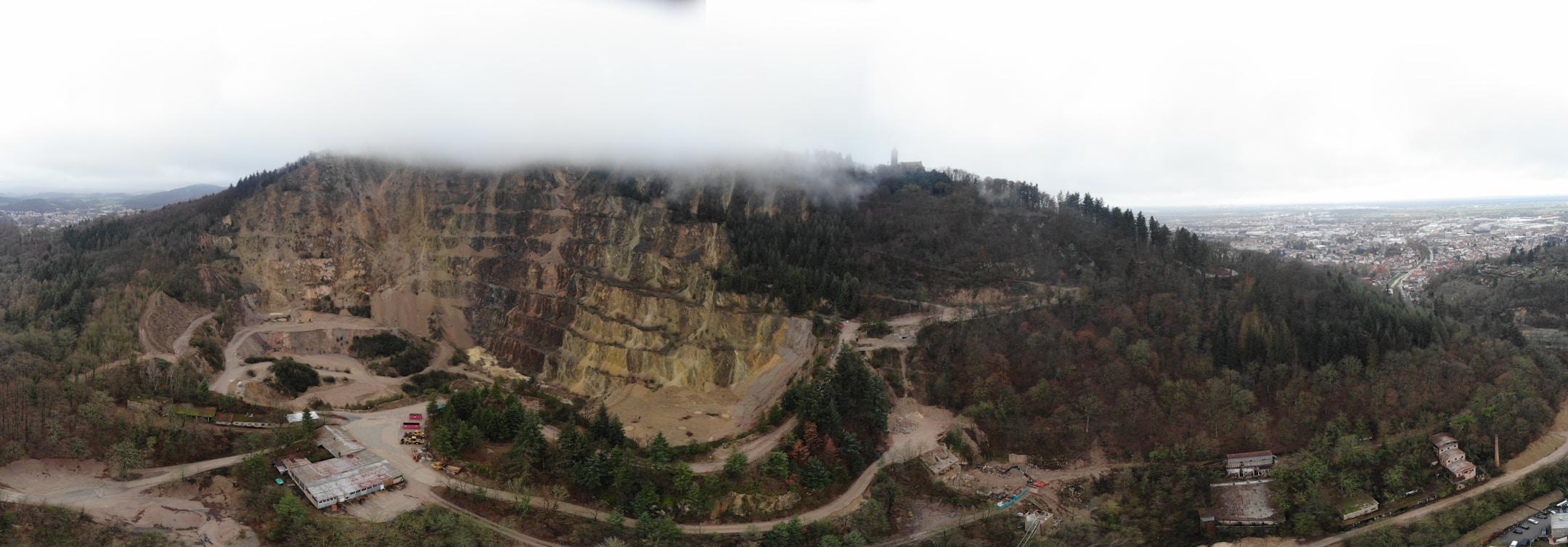 This panoramic image shows a sweeping view of a large quarry with exposed earth and sections of vegetation. Low clouds or fog partially obscure the top of the area, hinting at a high elevation or hilly terrain. In the distance, one can see a dense urban area, indicating the proximity of the quarry to a city or town. The contrast between the man-made excavation site and the natural, more lush surroundings is striking, as is the contrast between the rural and urban areas