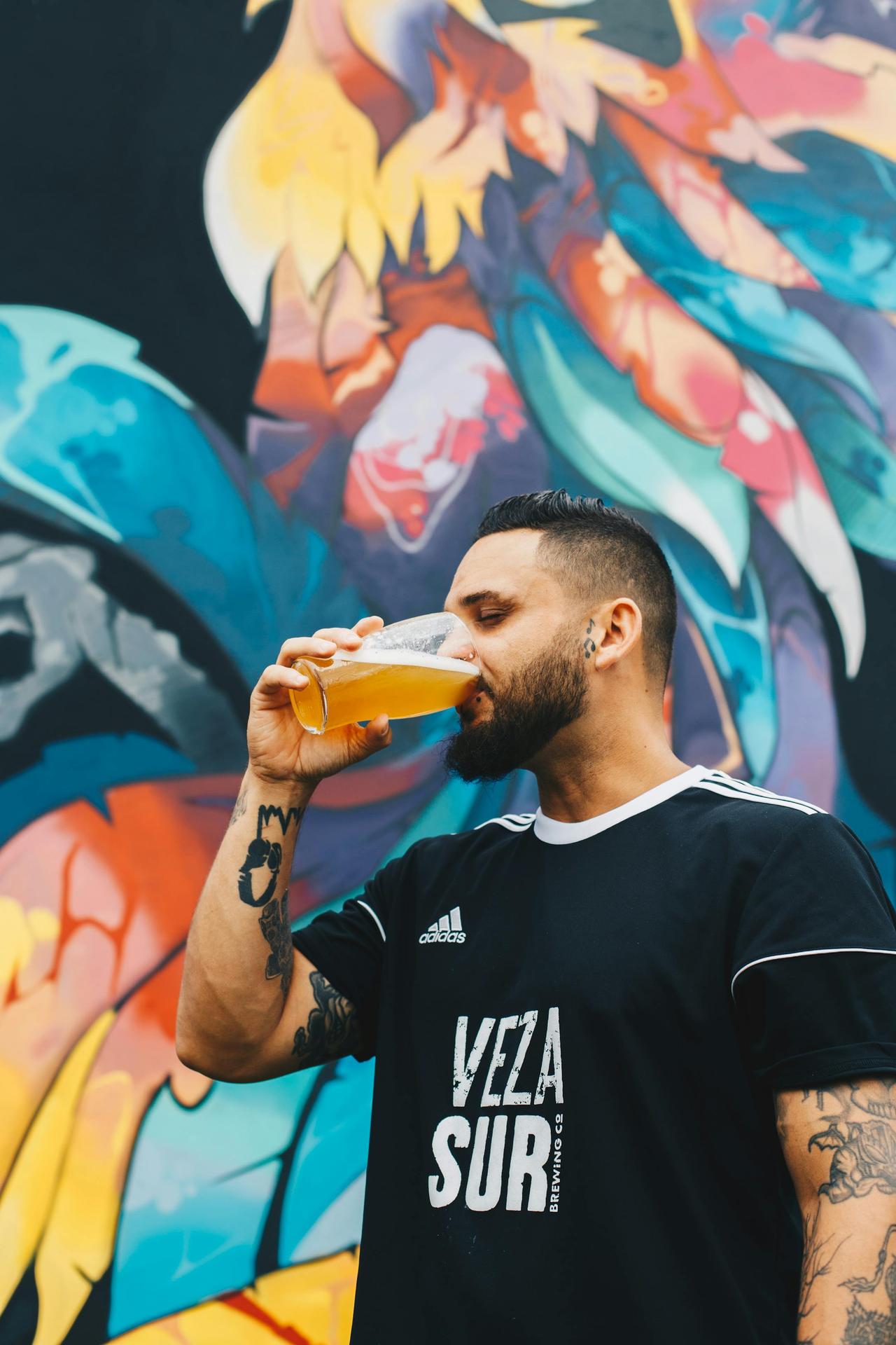 Man with tattoos enjoying a cold beer against a vibrant graffiti backdrop, embodying urban lifestyle.