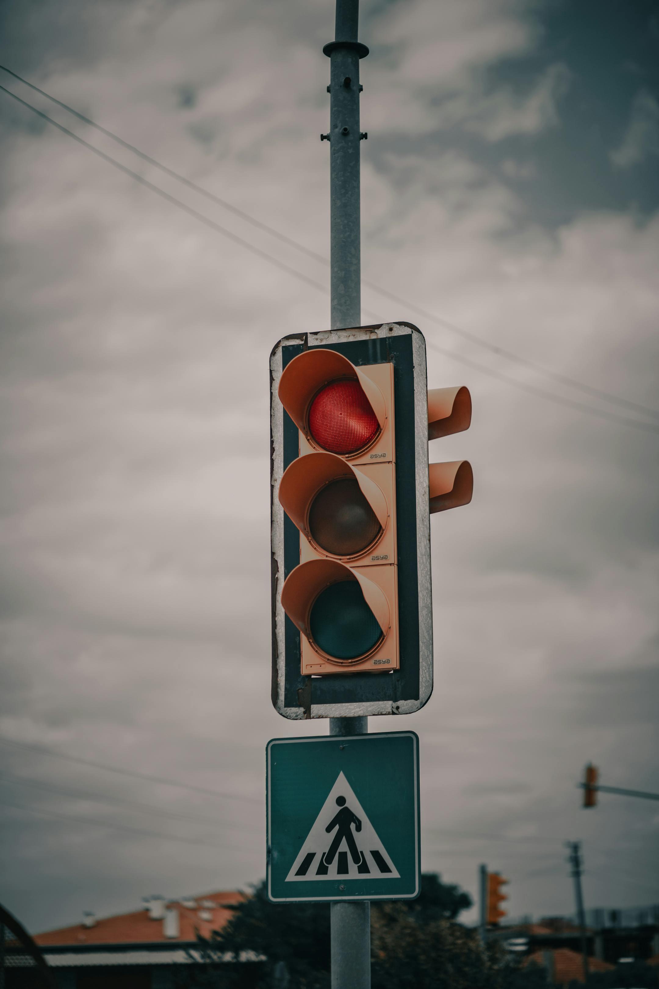 A red traffic light with a pedestrian sign, set under an overcast sky, signaling caution on the road.