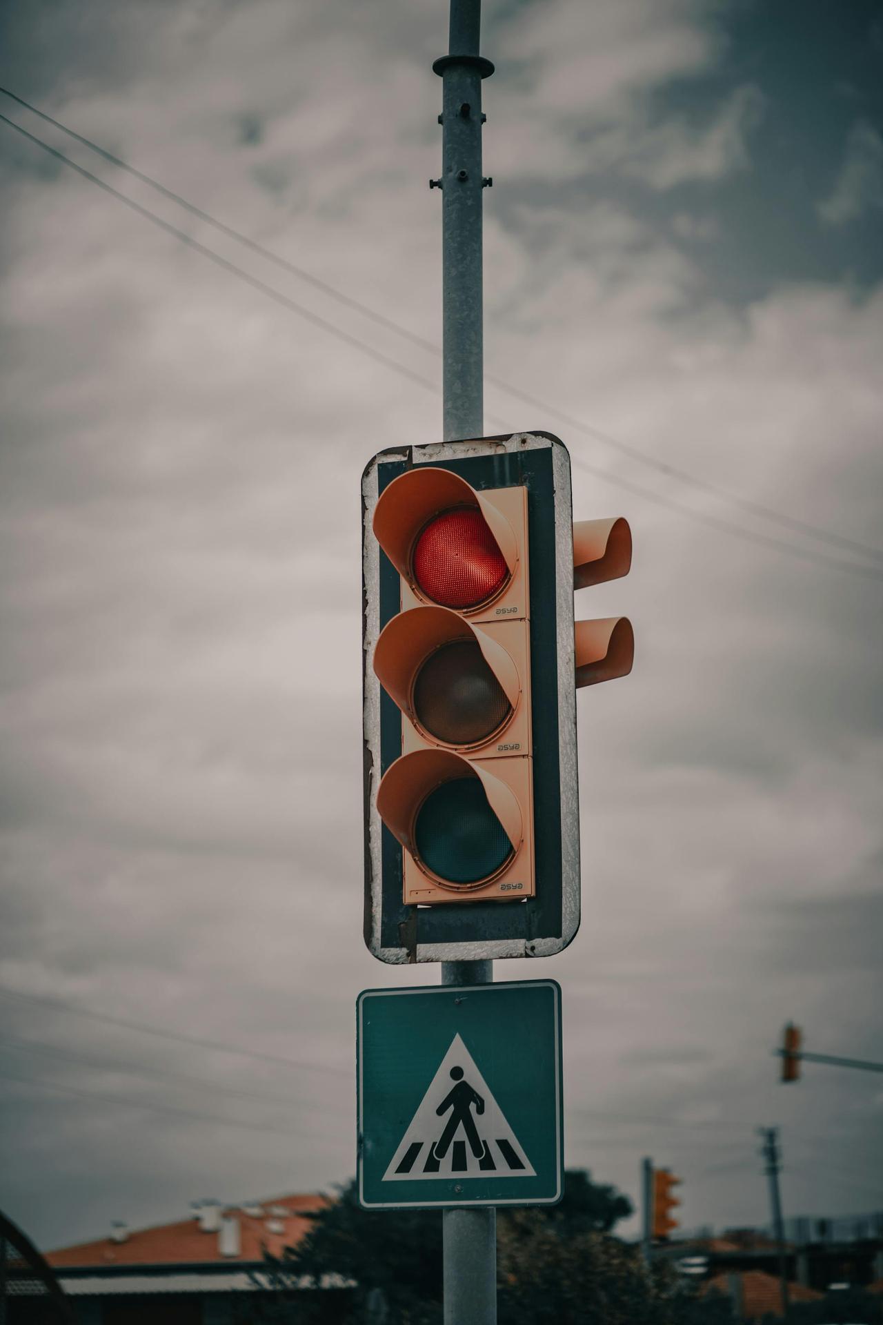 A red traffic light with a pedestrian sign, set under an overcast sky, signaling caution on the road.