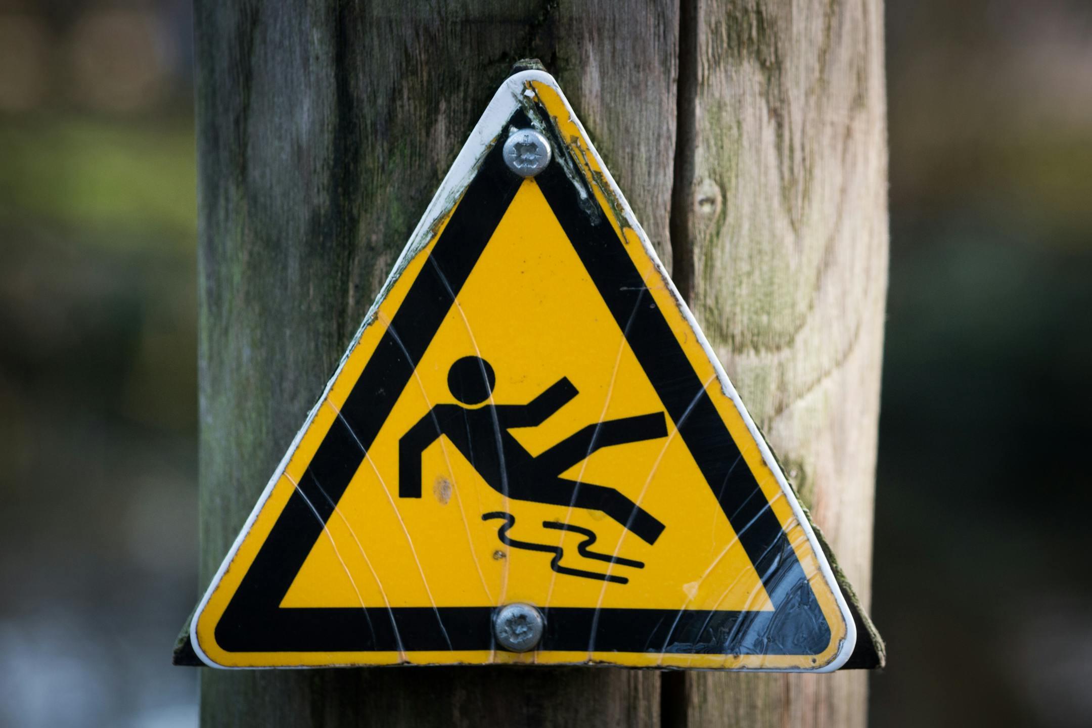 Close-up of a triangular warning sign indicating a slippery surface, fixed to a wooden post.