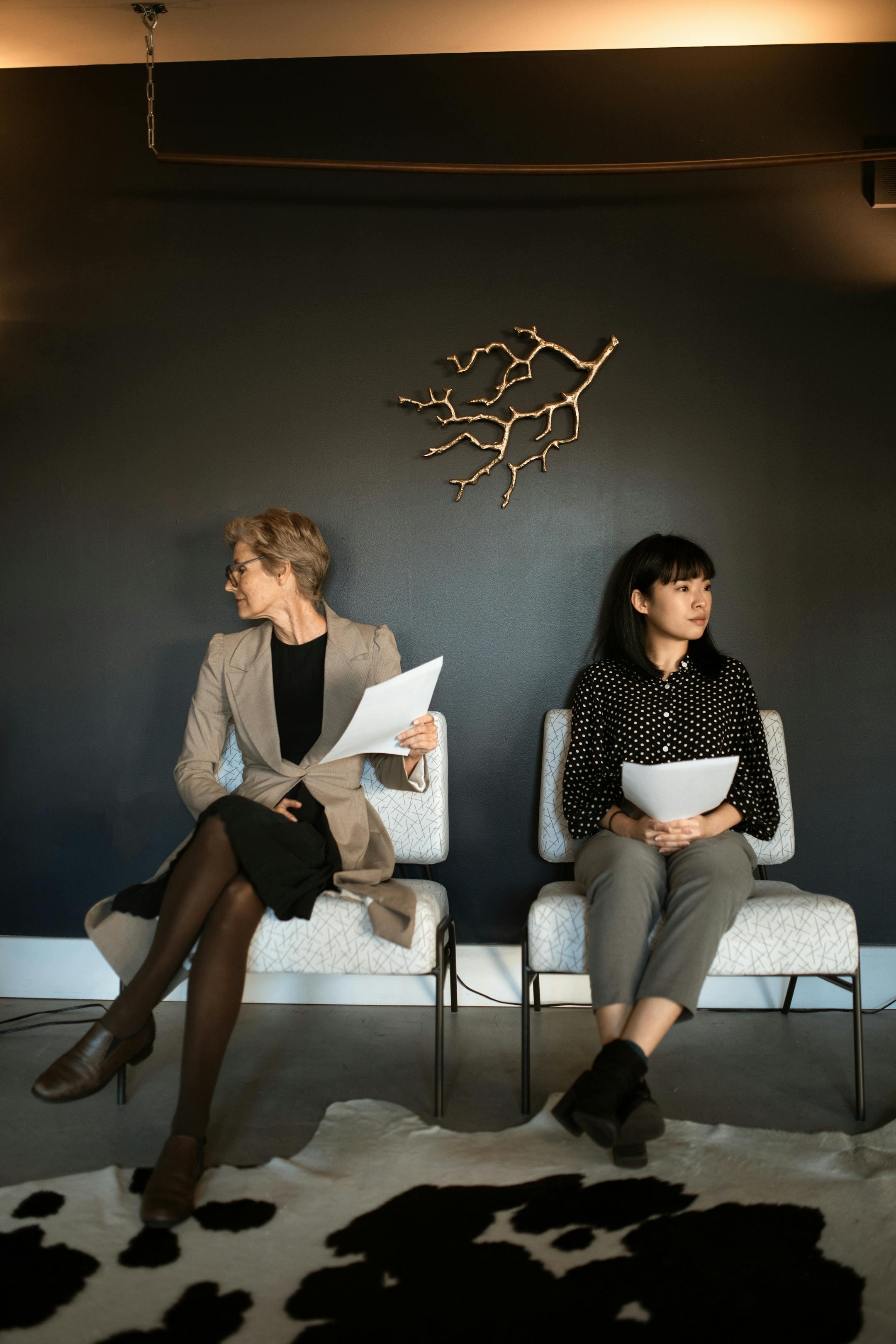 Two professional women seated in a modern office waiting area, holding documents in hand.