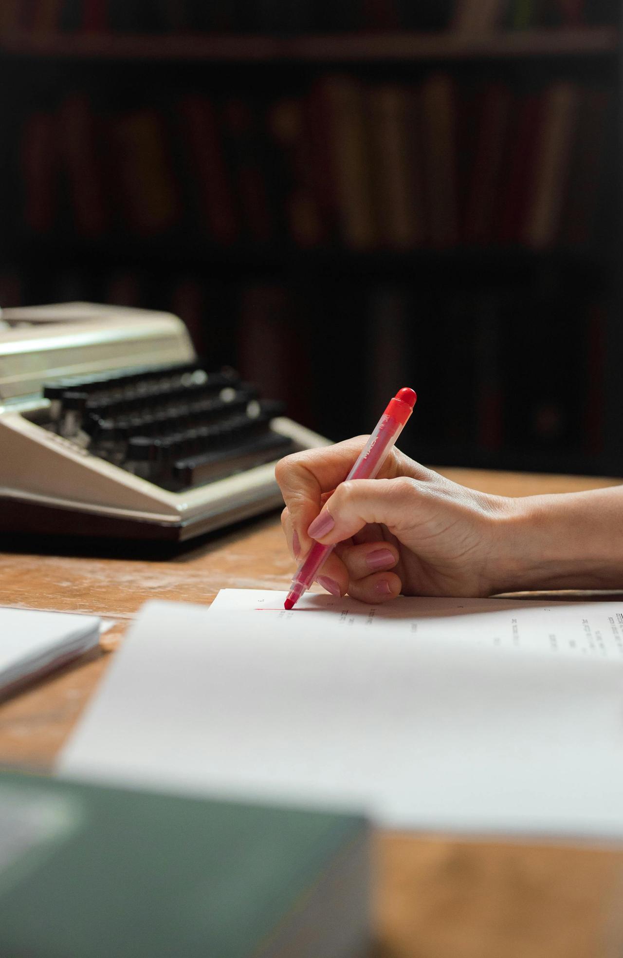 A close-up shot of a hand writing on papers with a red pen beside a vintage typewriter.