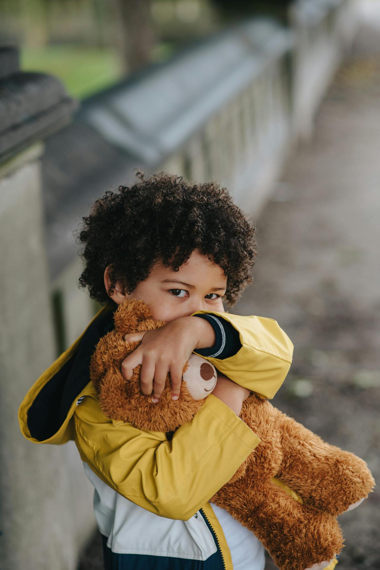 A cute child in a yellow jacket hugs a teddy bear with a blurred background.