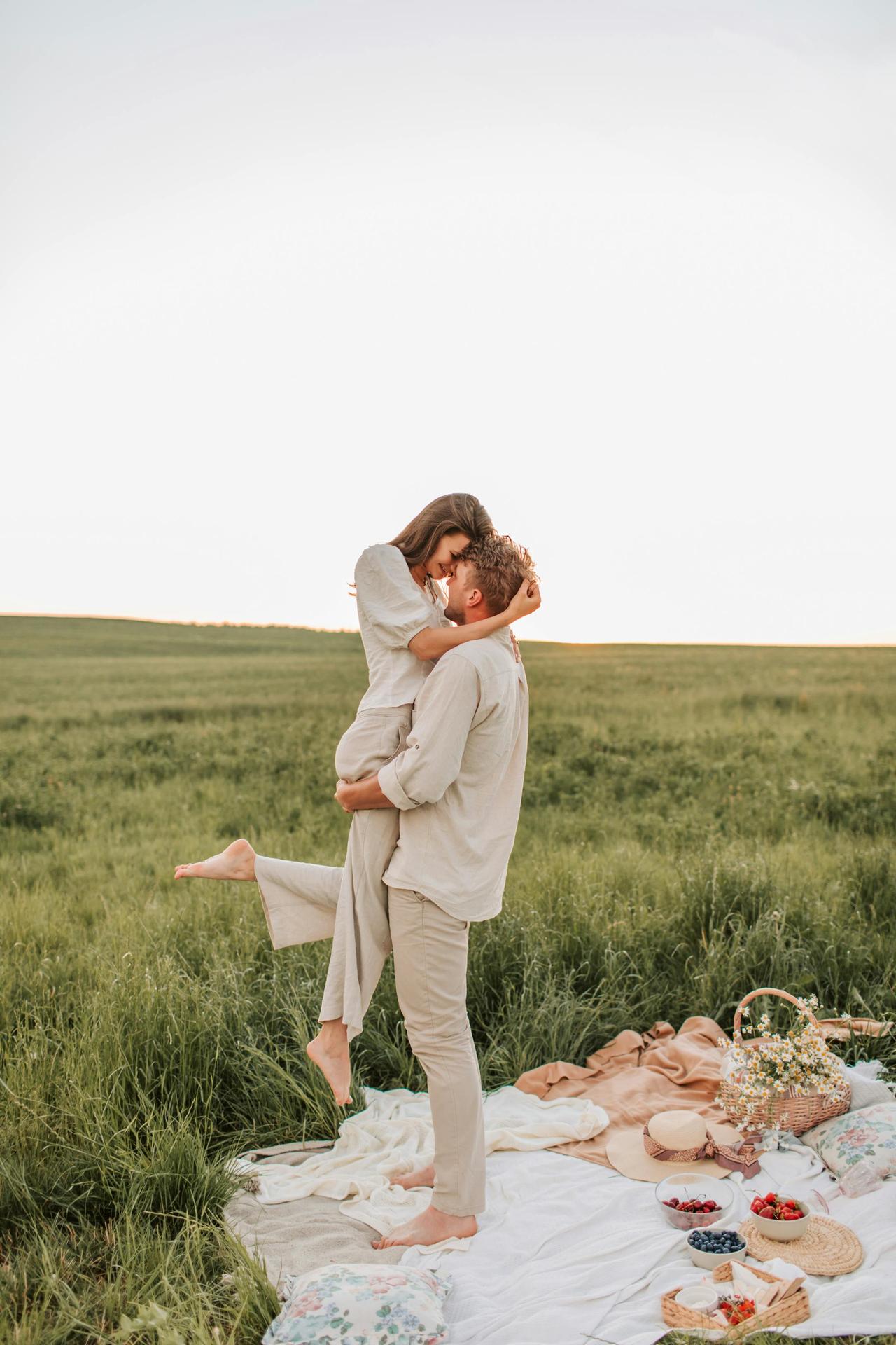 Couple enjoying a romantic picnic in a lush green field during sunset with a warm, loving embrace.