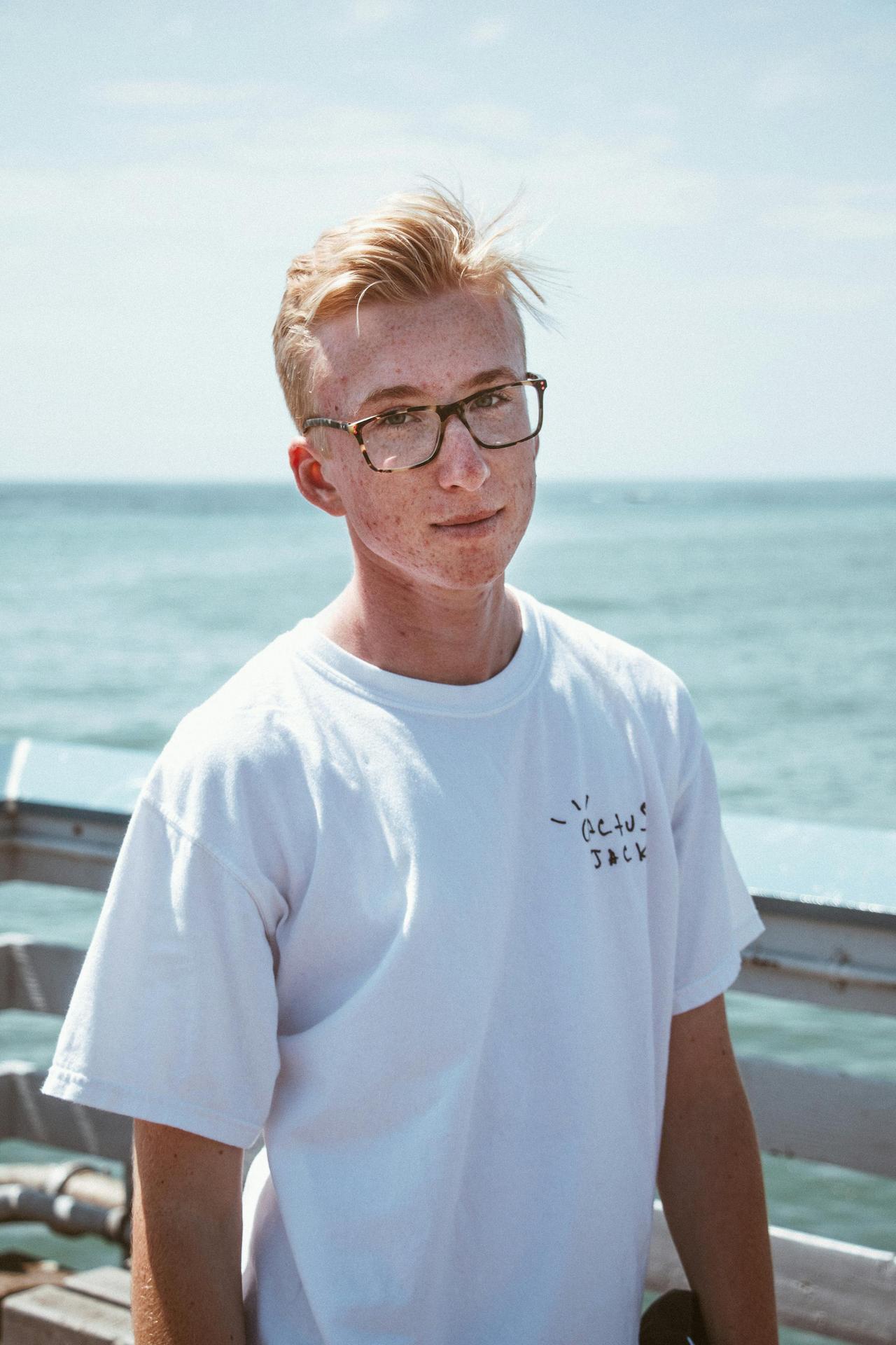 Young man in glasses wearing a white shirt, standing by the sea on a sunny day.