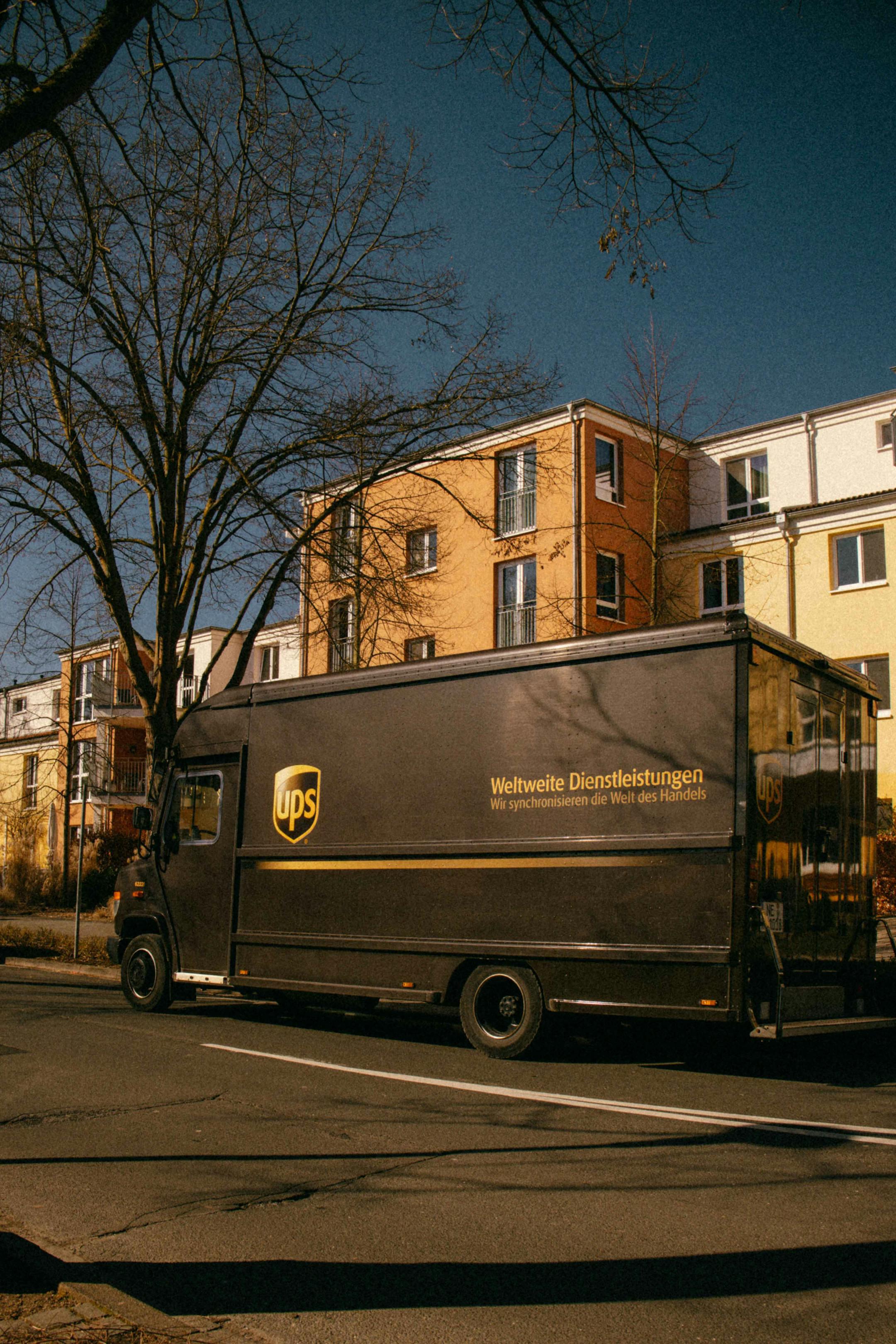 A UPS truck parked on a sunny urban street with apartment buildings in the background.