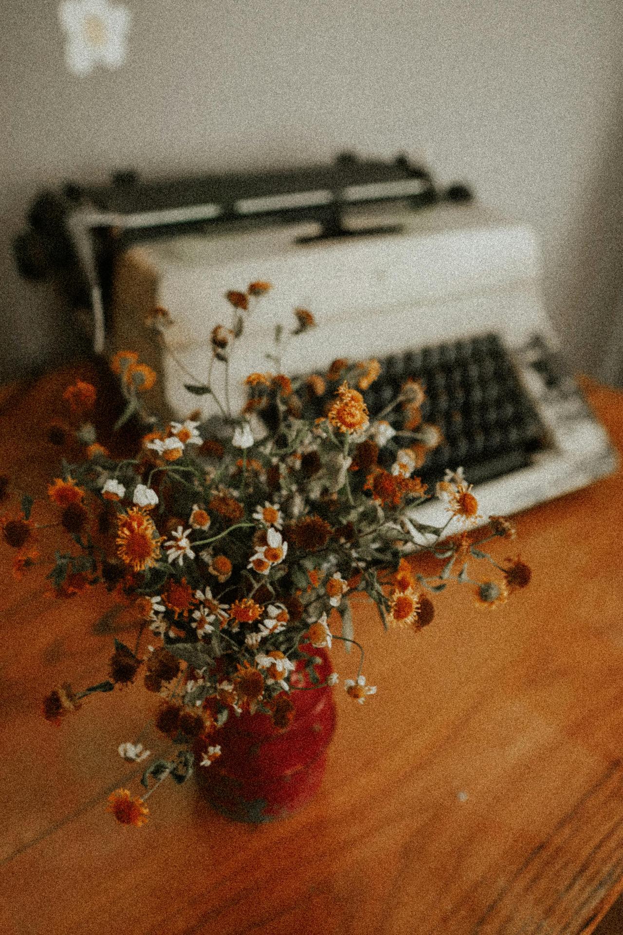 Aesthetic vintage setup featuring a typewriter and dried flowers in a red vase on a wooden table.