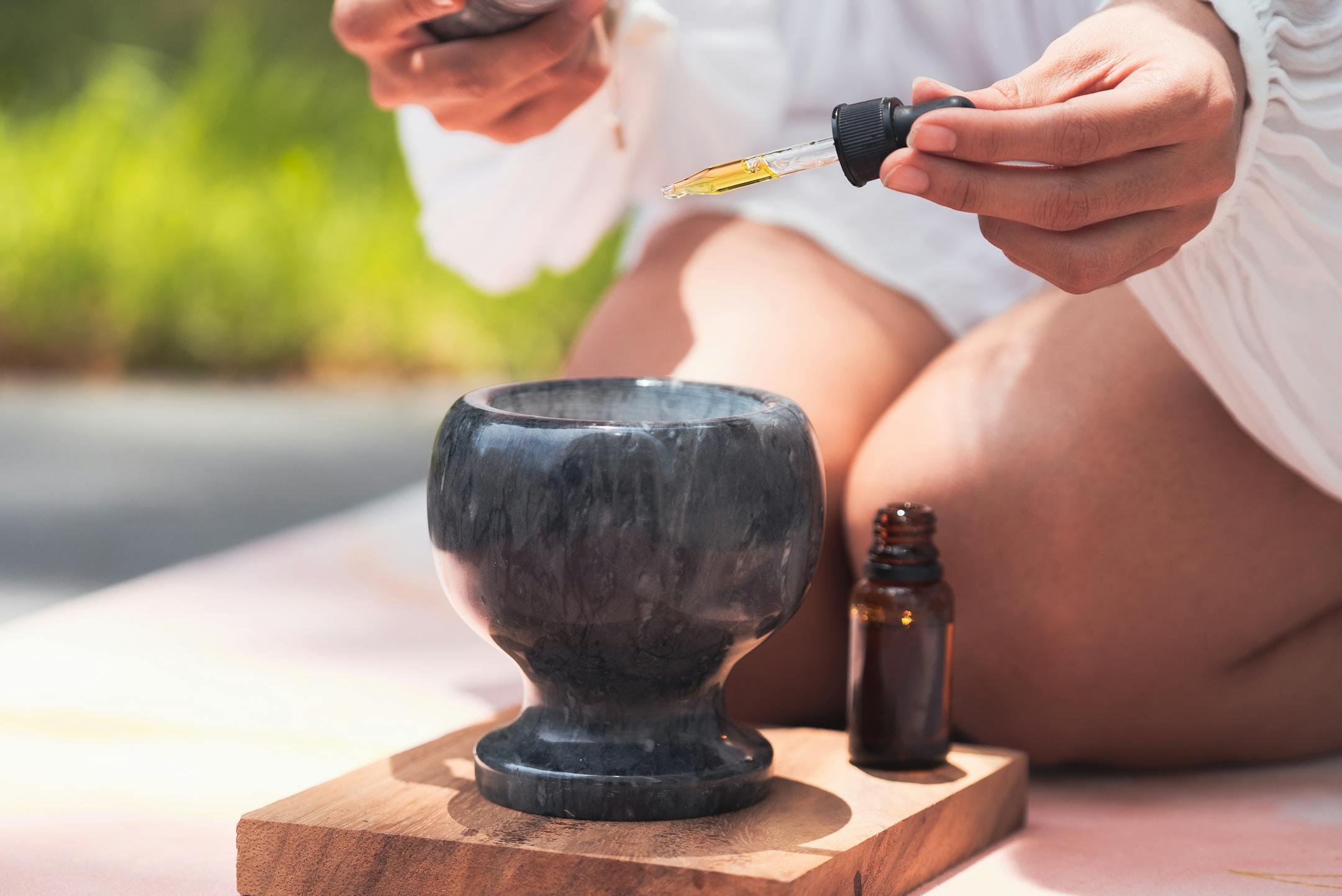 Close-up of a hand holding a dropper of essential oil above a ceramic bowl outdoors.