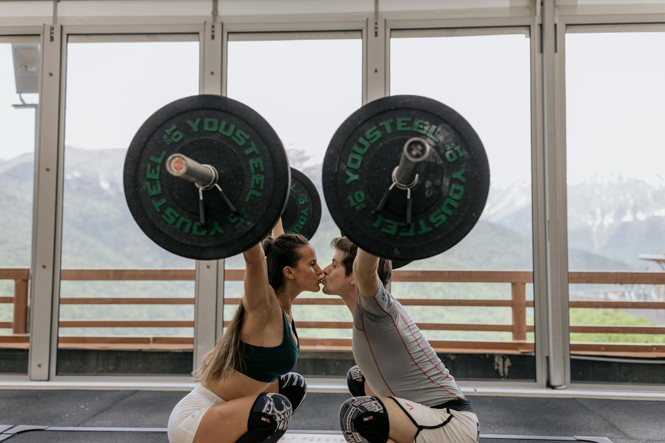 A couple in athletic gear kisses while lifting barbells in a modern gym setting.