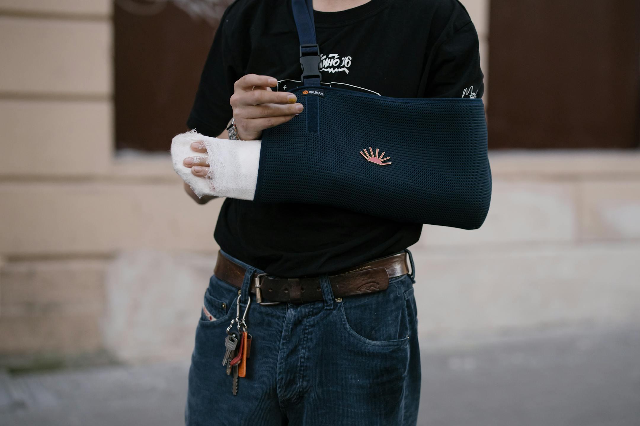 A man wearing a dark sling with a bandaged arm, standing outdoors in casual attire.