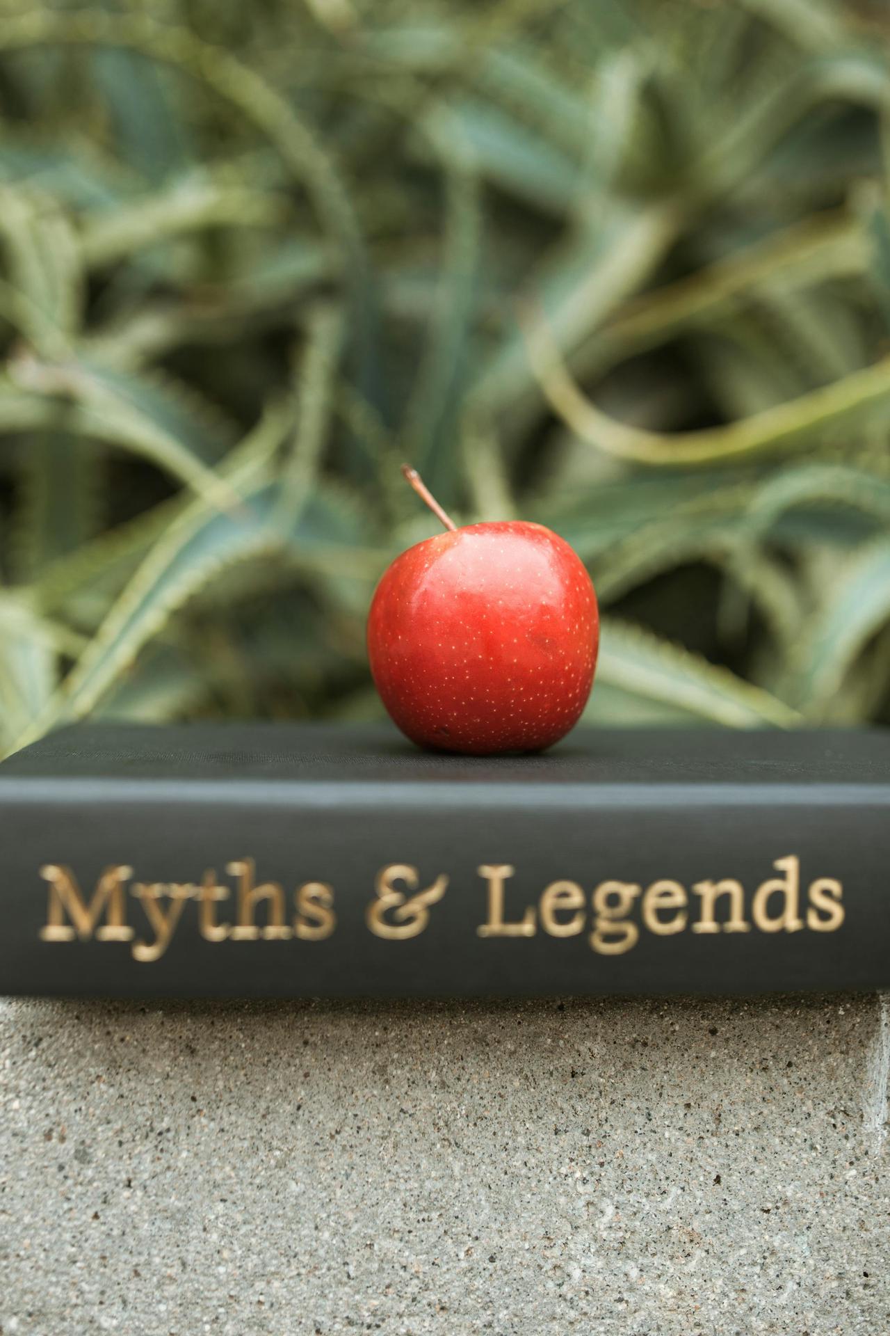 Red apple placed atop a 'Myths & Legends' book against an abstract background, representing knowledge and temptation.