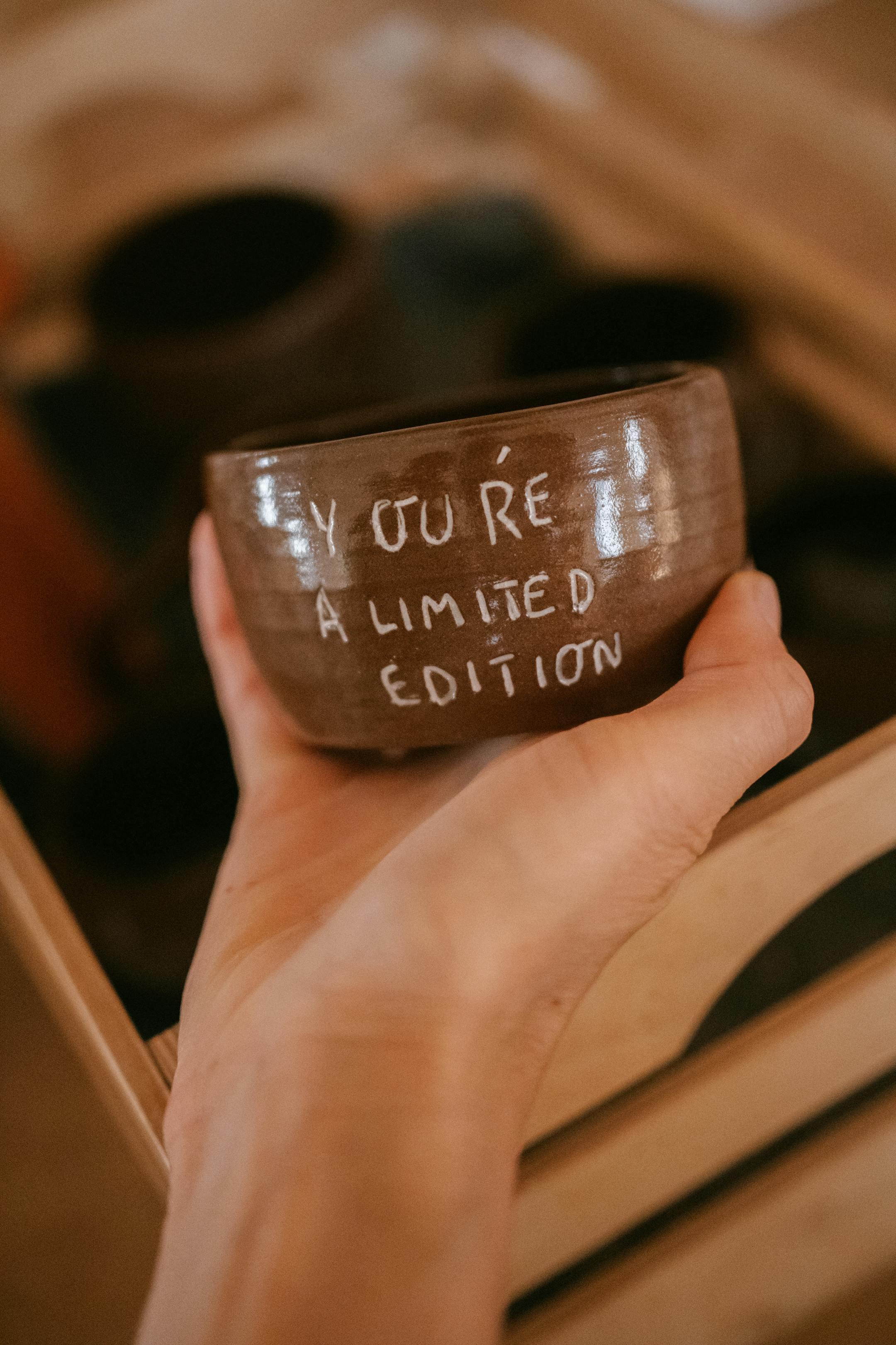 Close-up of a hand holding a ceramic bowl with 'You're a limited edition' text, indoors.