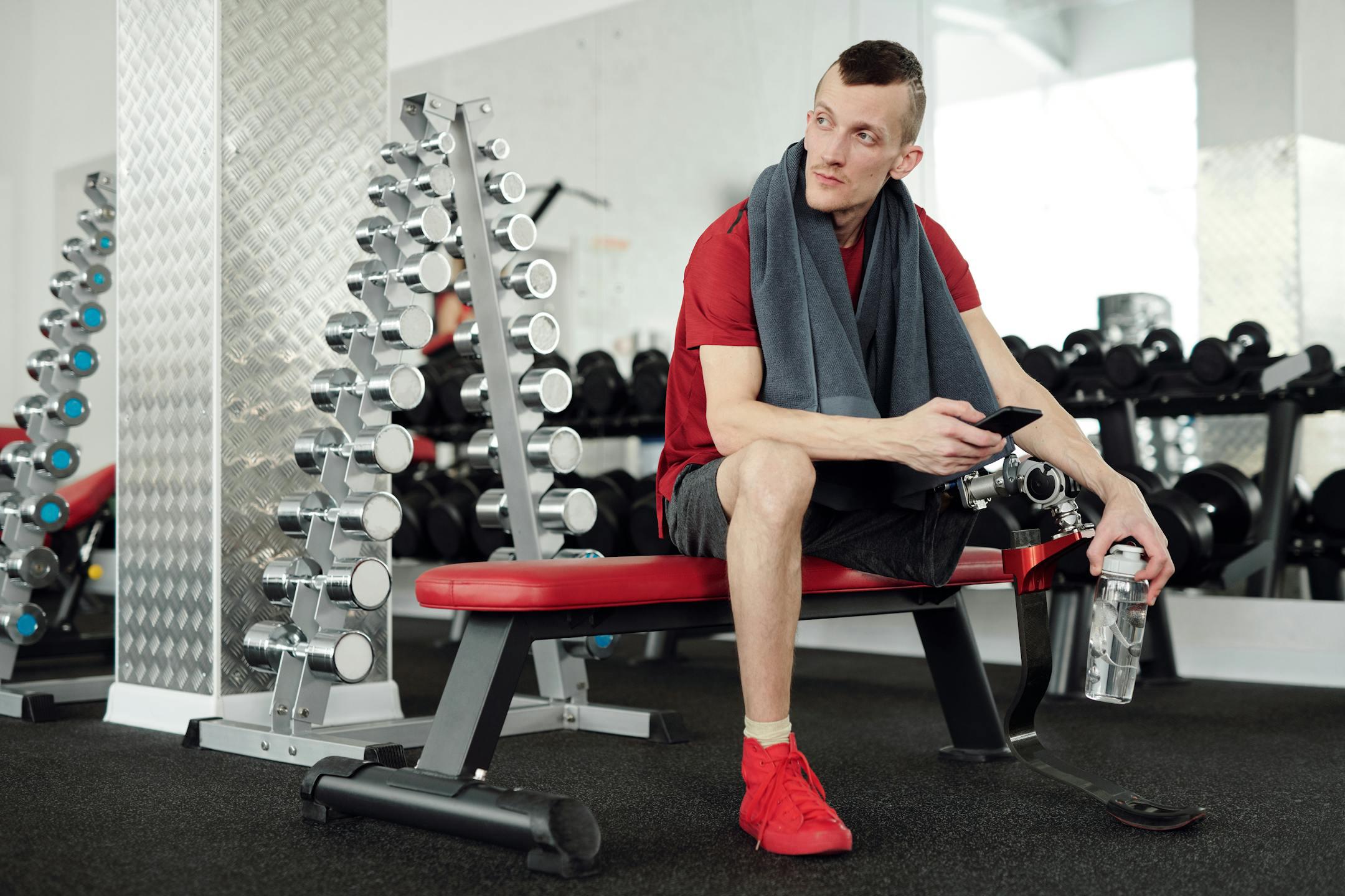 Man with prosthetic leg taking a break in gym, holding smartphone and water bottle.