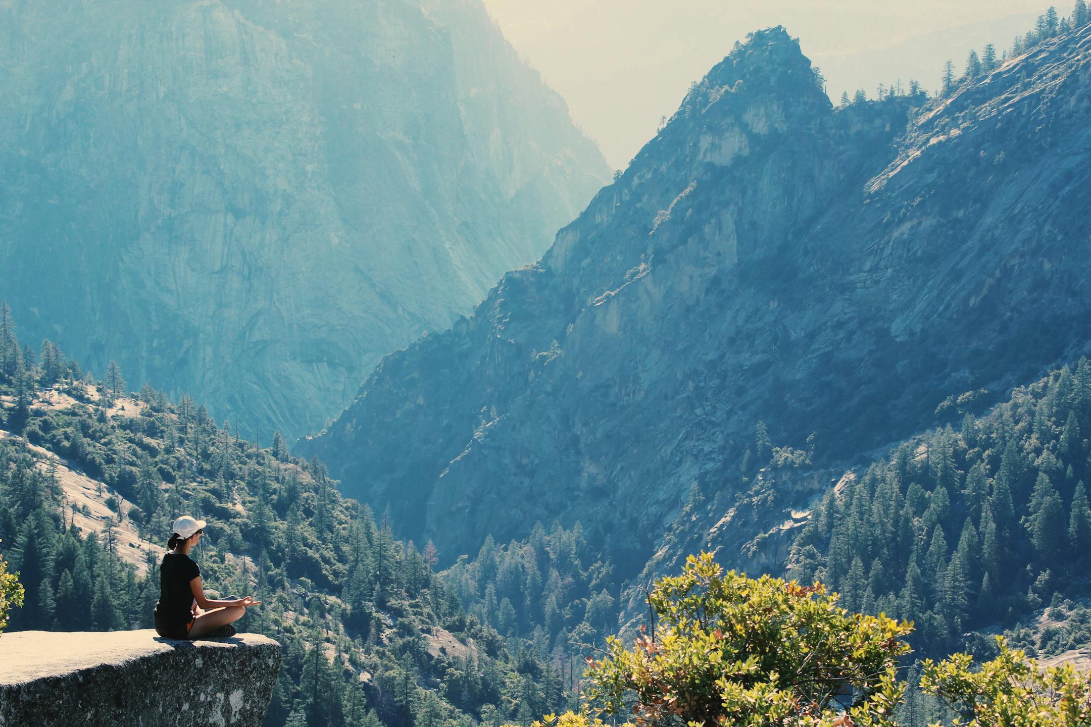A woman practices meditation on a cliff edge, surrounded by majestic mountains and serene nature.