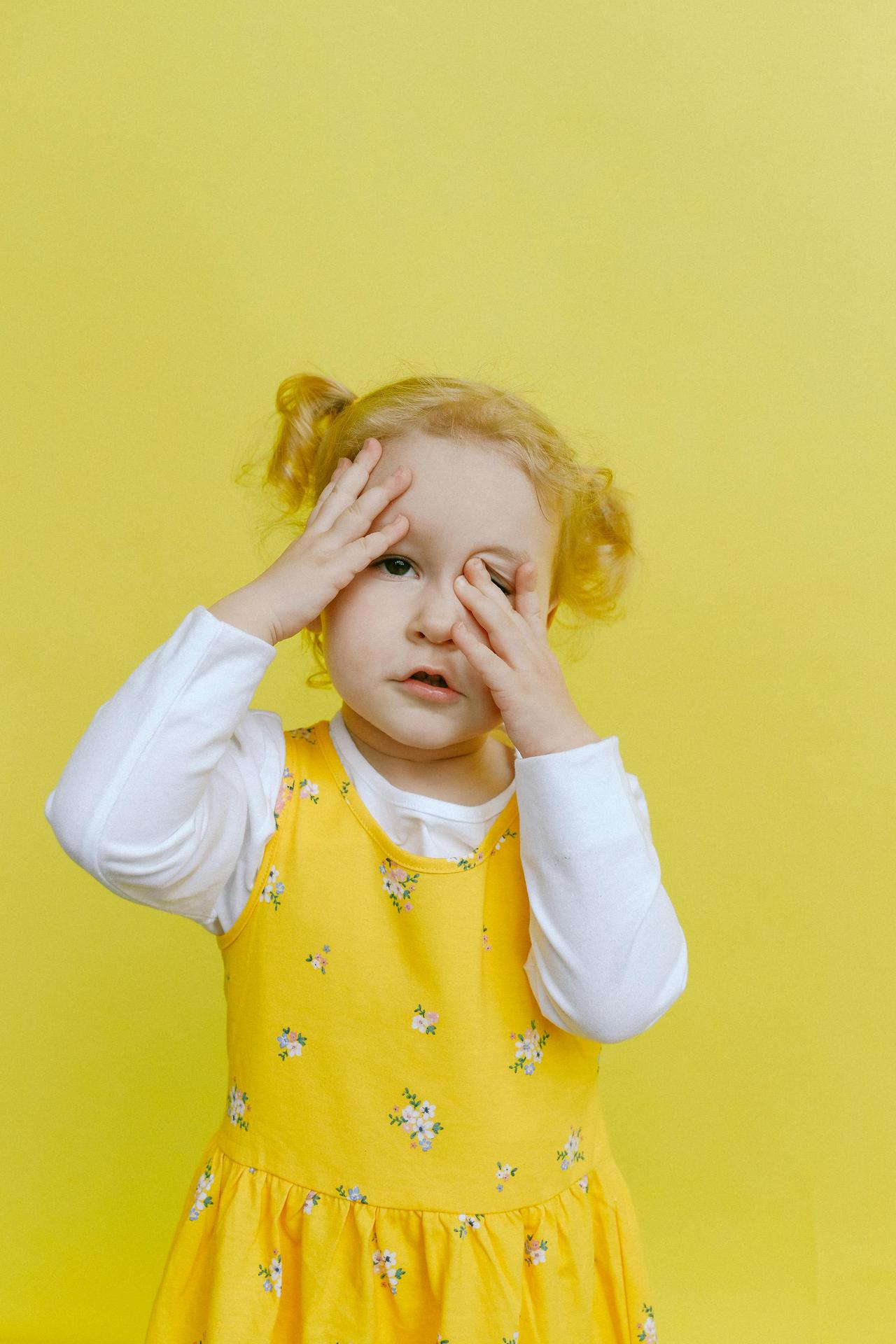 Adorable toddler with hands on face wearing a yellow floral dress against a yellow background.