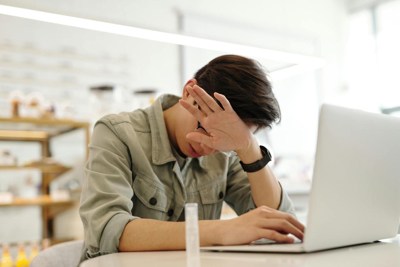 An adult man appears stressed while working on a laptop indoors, covering his face with one hand.