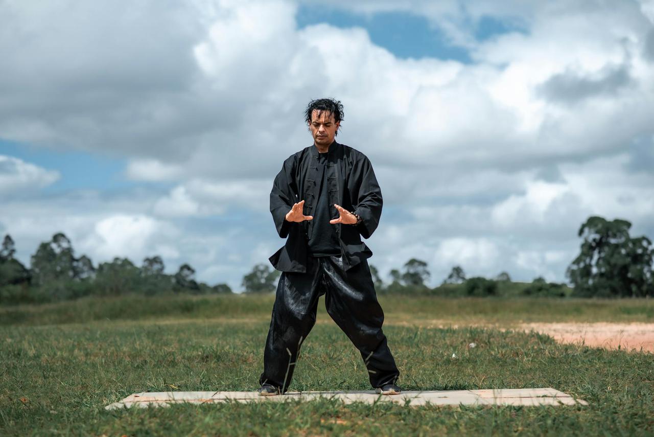 Man practicing martial arts outdoors in black uniform, performing hand gestures on a cloudy day.