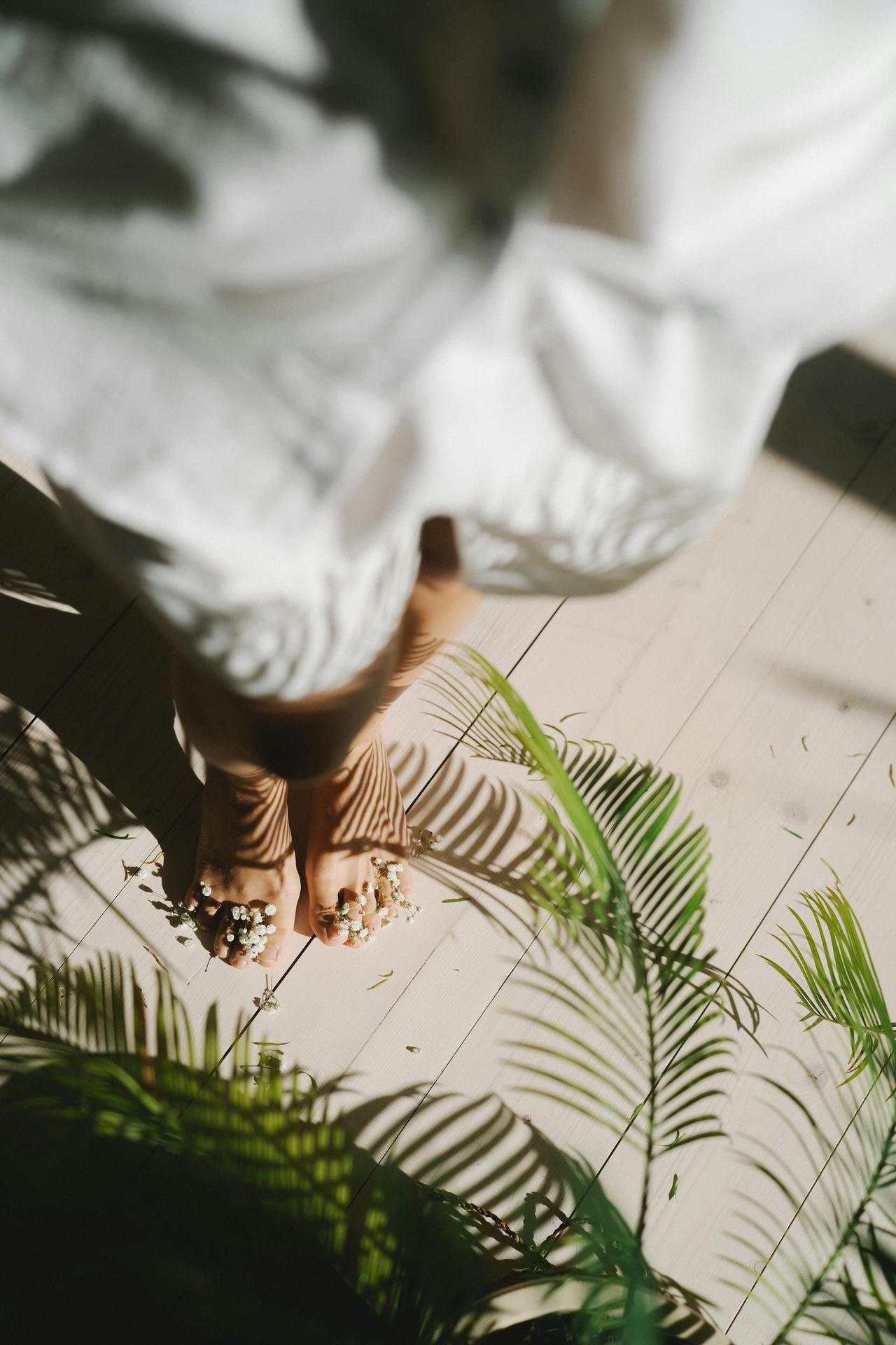A high angle artistic photo of a person standing barefoot on a floor beneath leafy shadows.